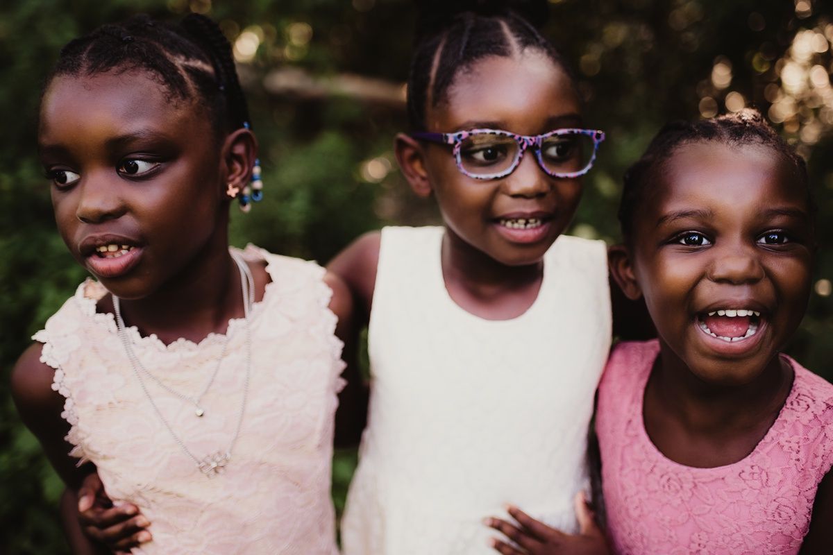 Three children wearing dresses smile at the camera with one wearing glasses in the middle.