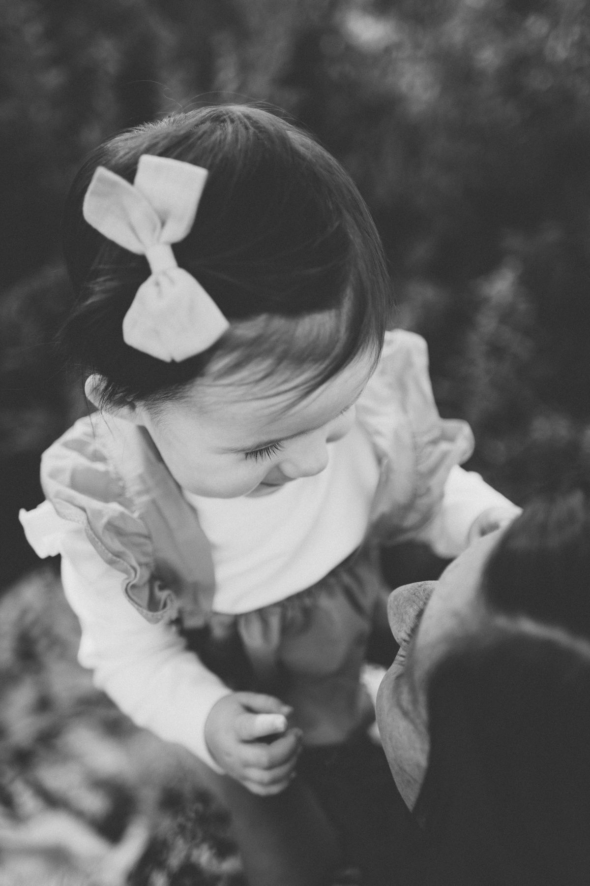 A black and white portrait of a child with a bow being held in nature.