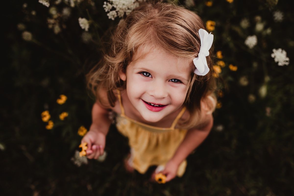 Close-up outdoor portrait of a child wearing a yellow dress and white bow among flowers.