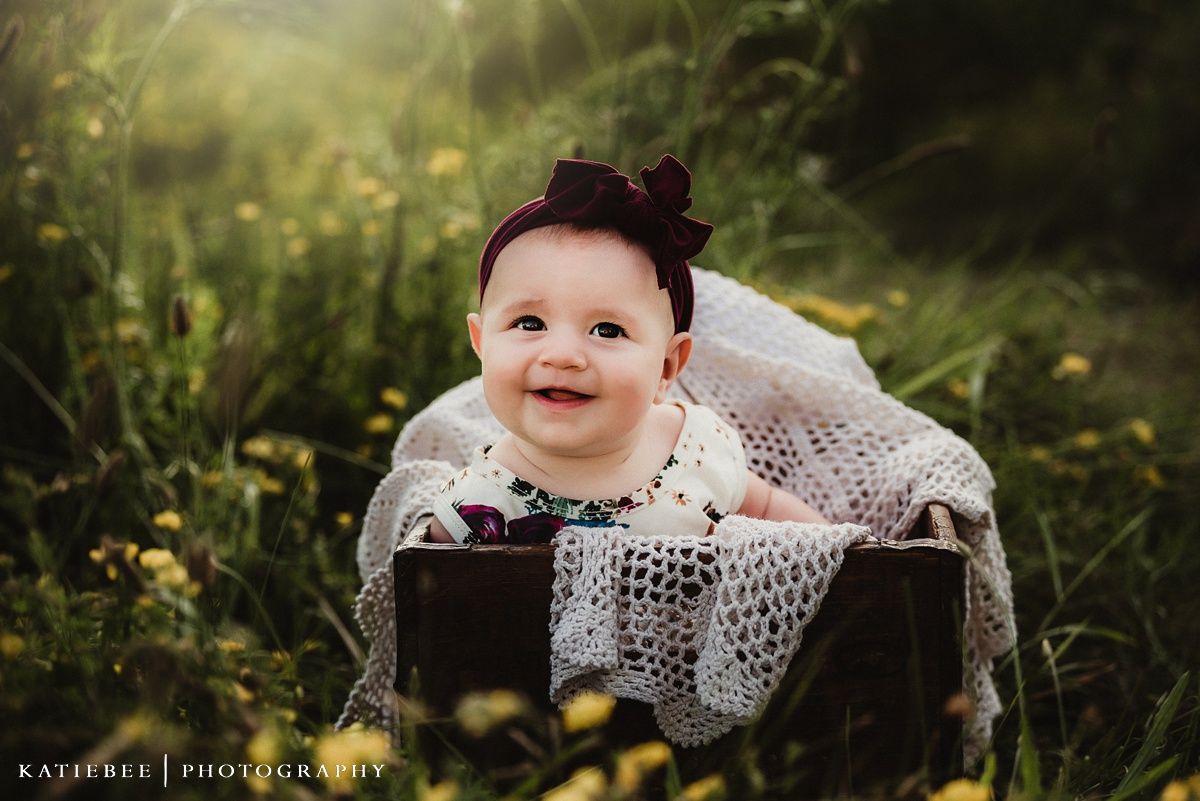 Happy baby wearing white lace dress and burgundy bow sits in wooden box among wildflowers.