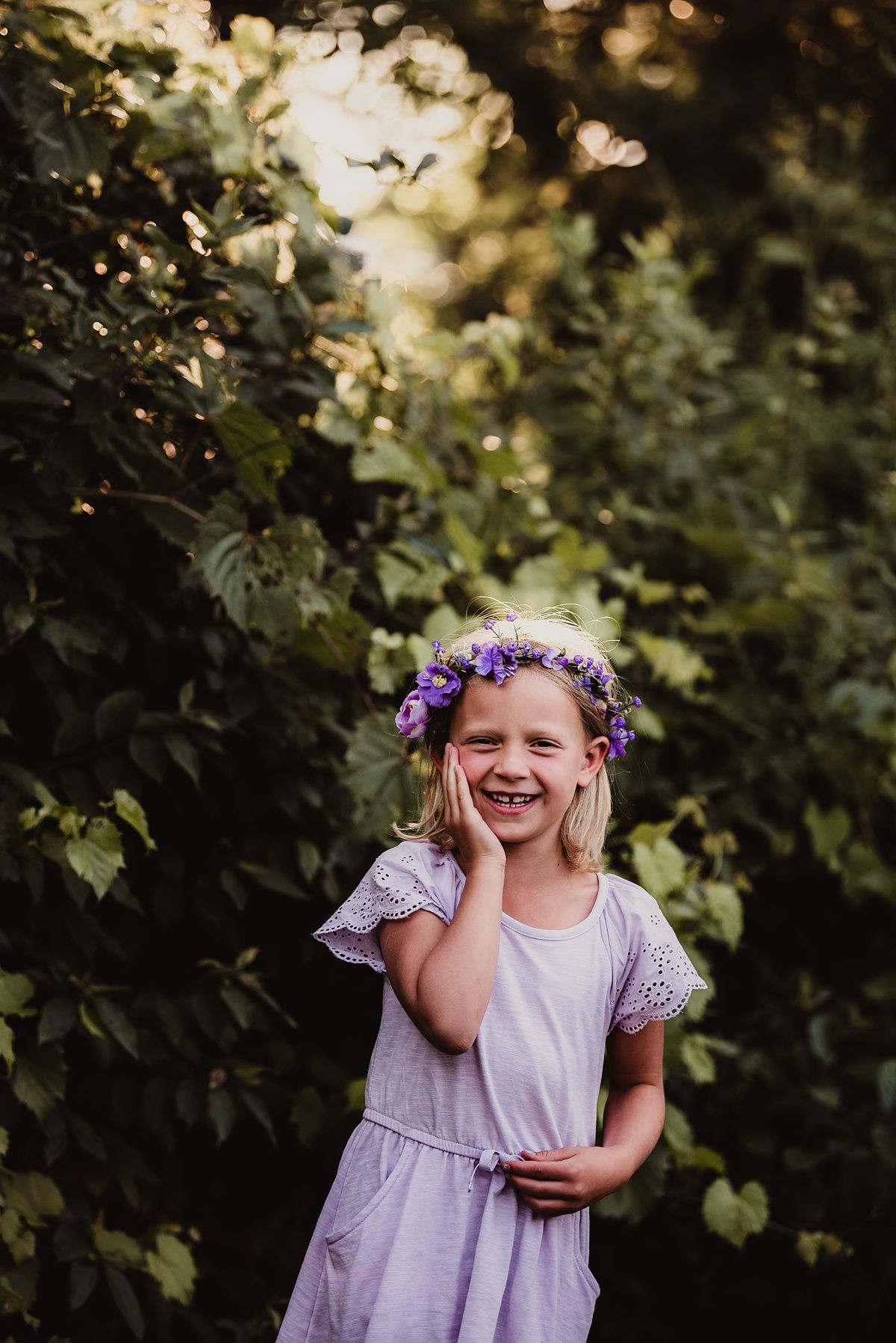 Someone in a lavender dress and flower crown smiling in a sunlit garden setting.