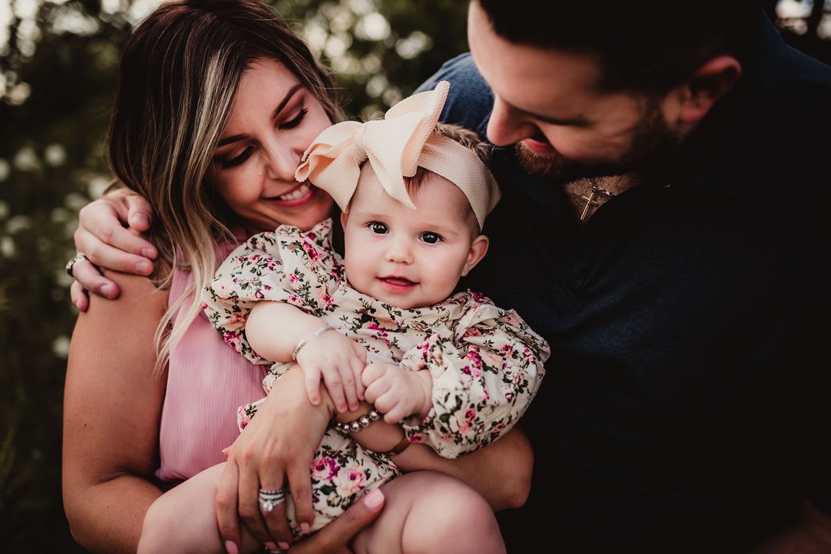 A family shares an intimate moment outdoors with their baby wearing a floral dress and bow.