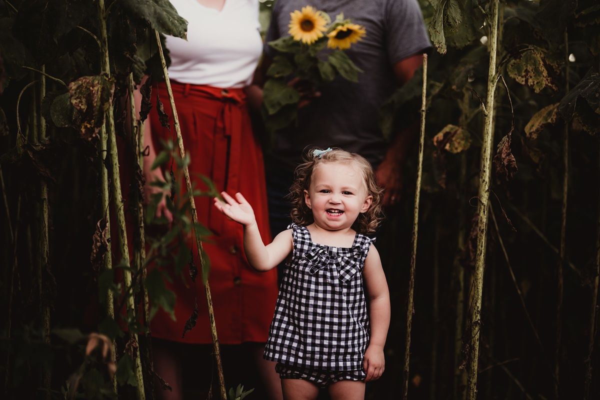 A child in a checkered dress waves while standing among tall sunflowers.