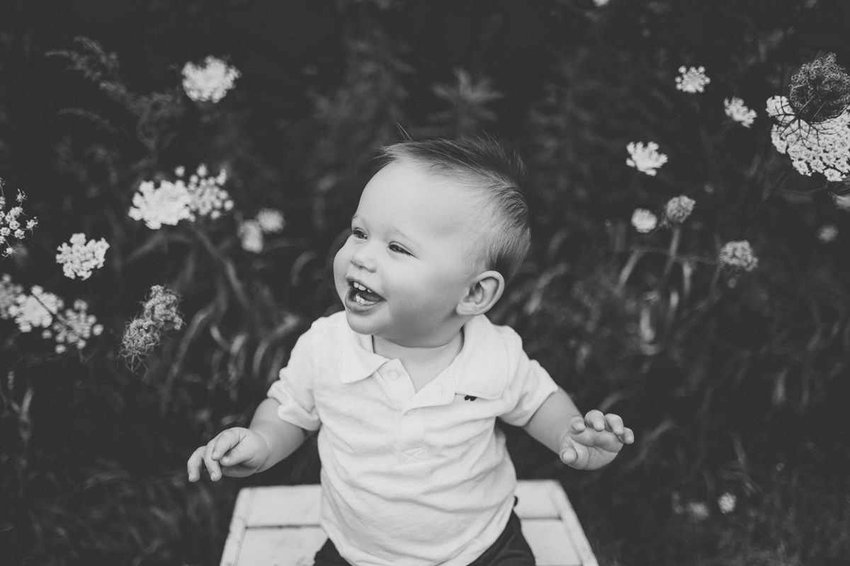 Black and white photo of a joyful baby sitting on a white wooden crate among wildflowers.