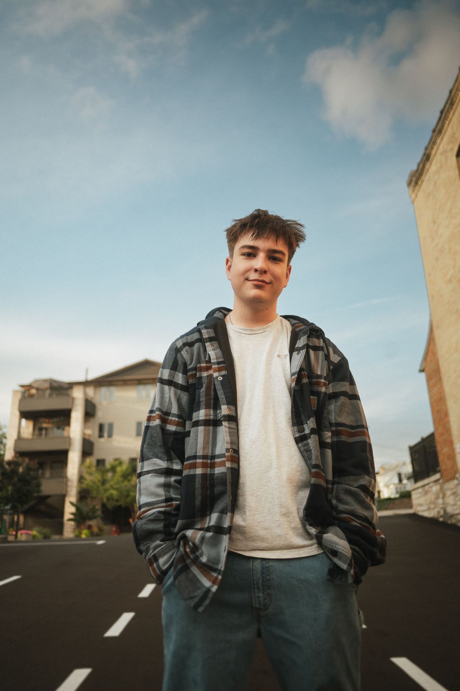 Person in plaid flannel jacket and jeans stands casually on an urban street with buildings in the background.