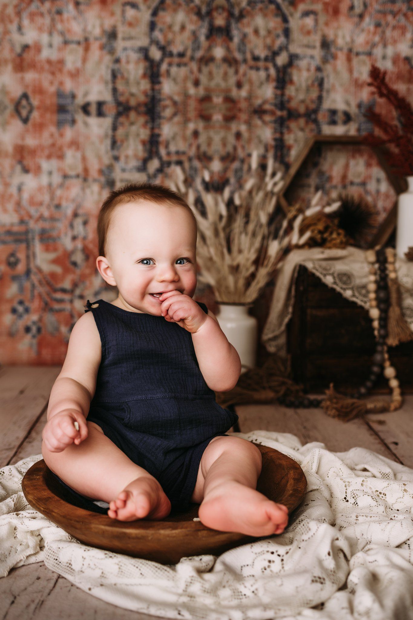 Baby wearing navy sleeveless outfit sits in wooden bowl with finger in mouth against patterned backdrop.