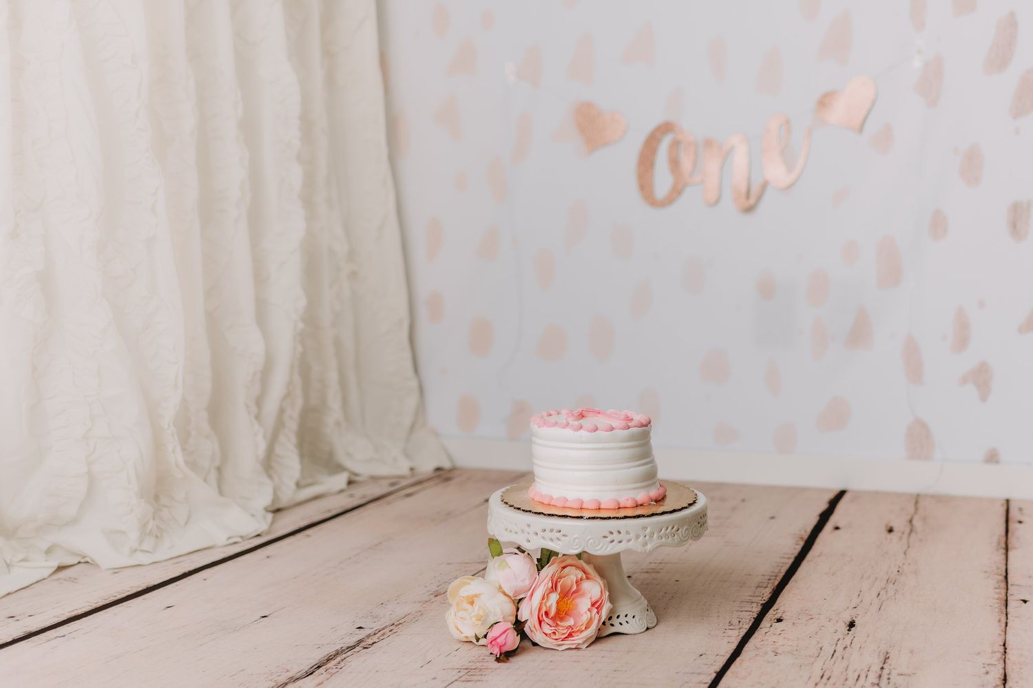Small pink and white cake on decorative stand with roses against light patterned background.