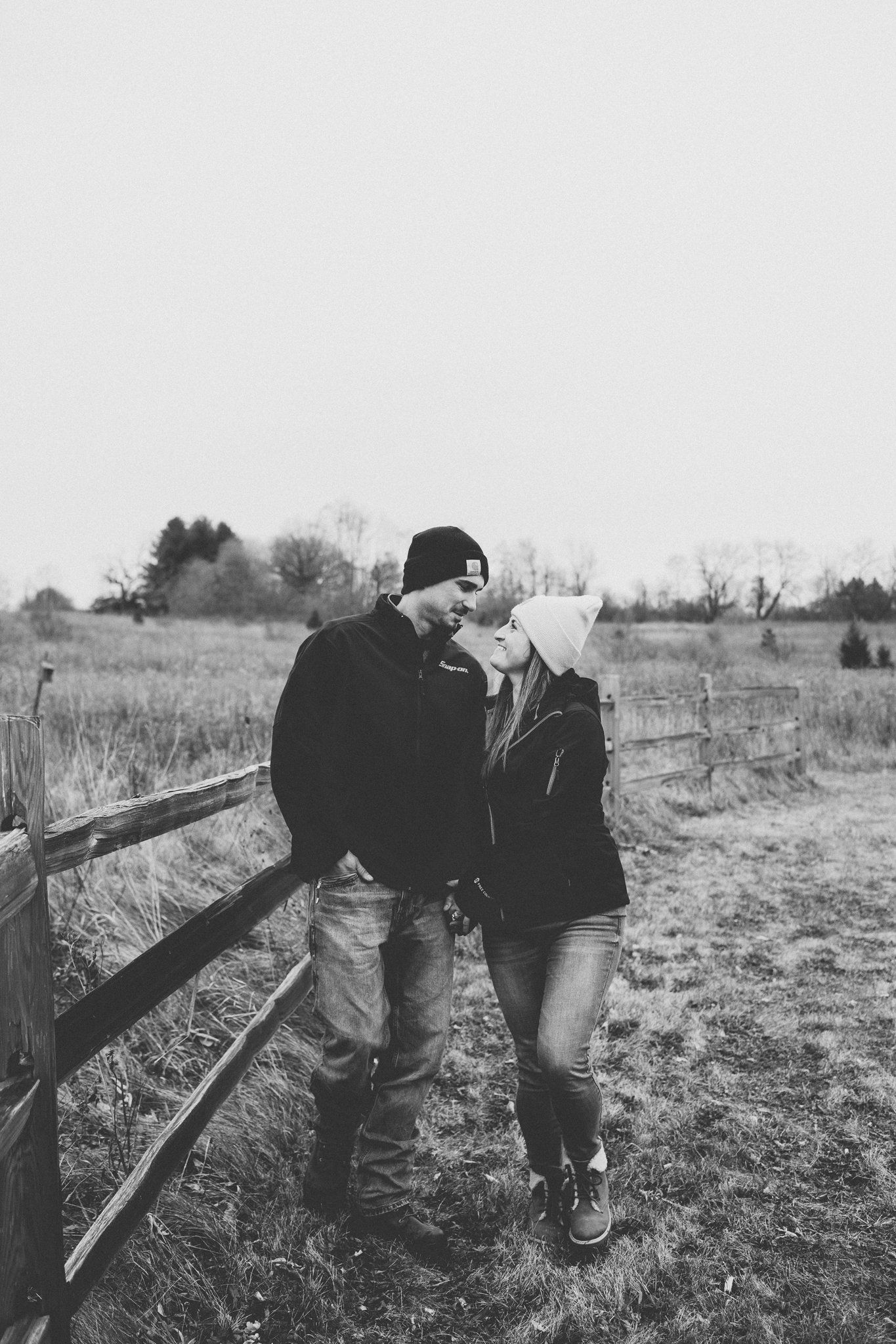 A couple in winter attire share a romantic moment while leaning against a wooden fence in a rural field.