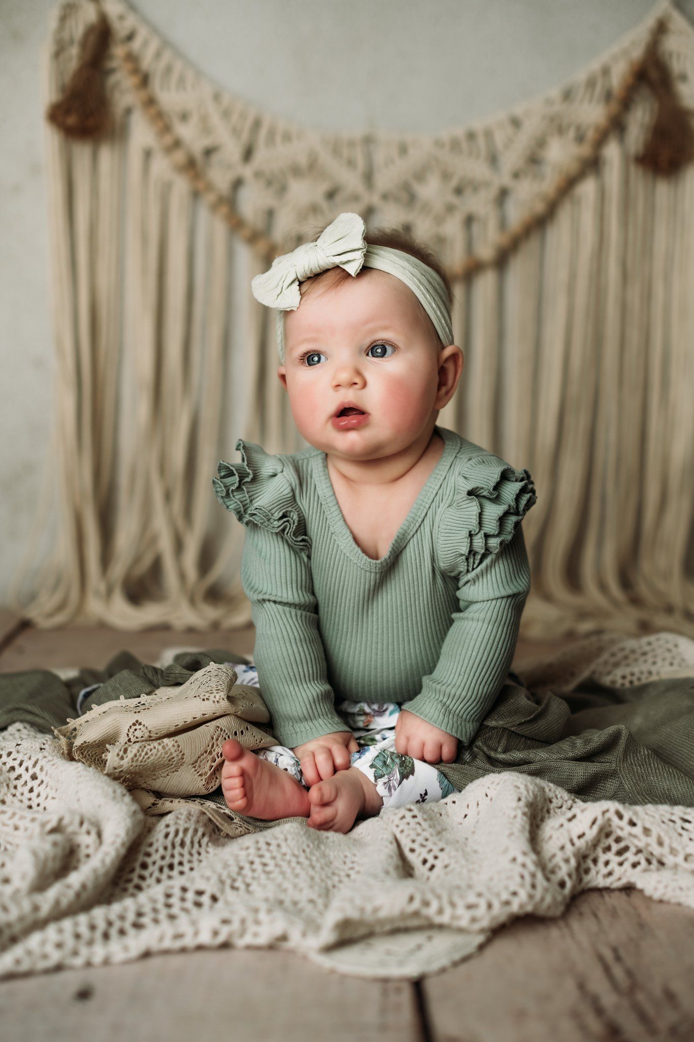 Baby in mint ruffled outfit sits upright on textured cream blanket against macrame backdrop.