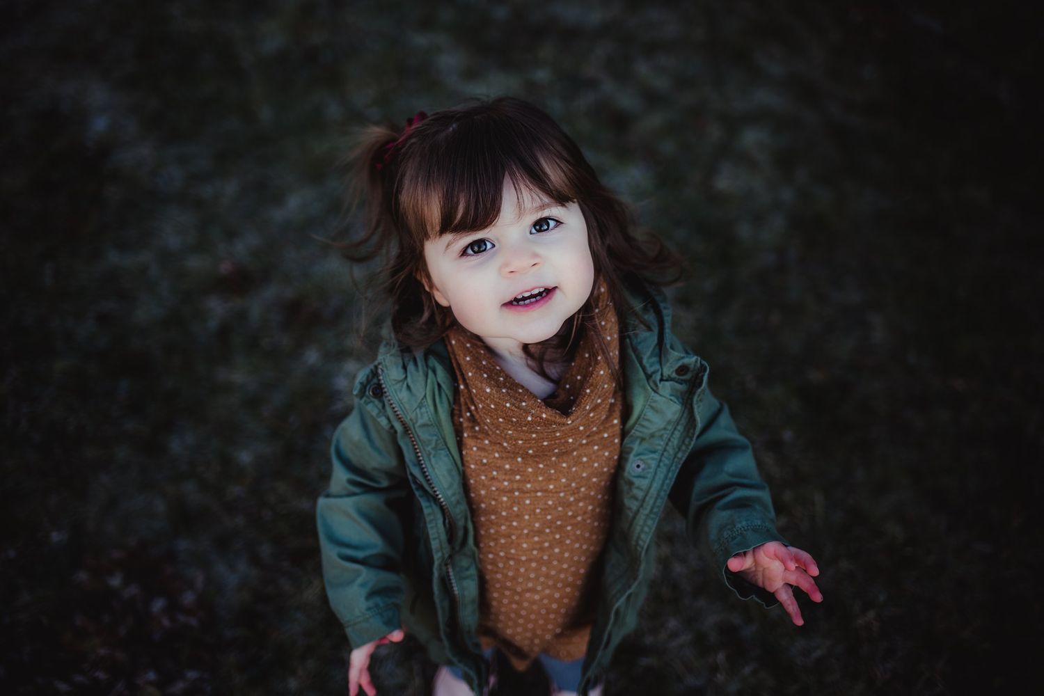 A child in a green jacket and polka dot dress poses outdoors on a dark evening.