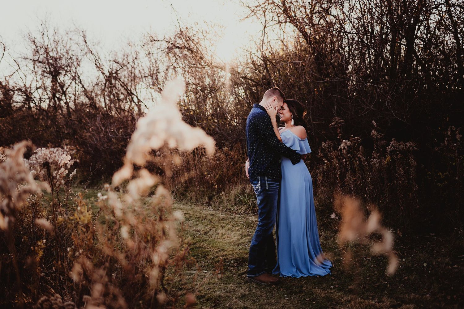A romantic sunset engagement photo shows a couple embracing in a field of dried flowers and brush.