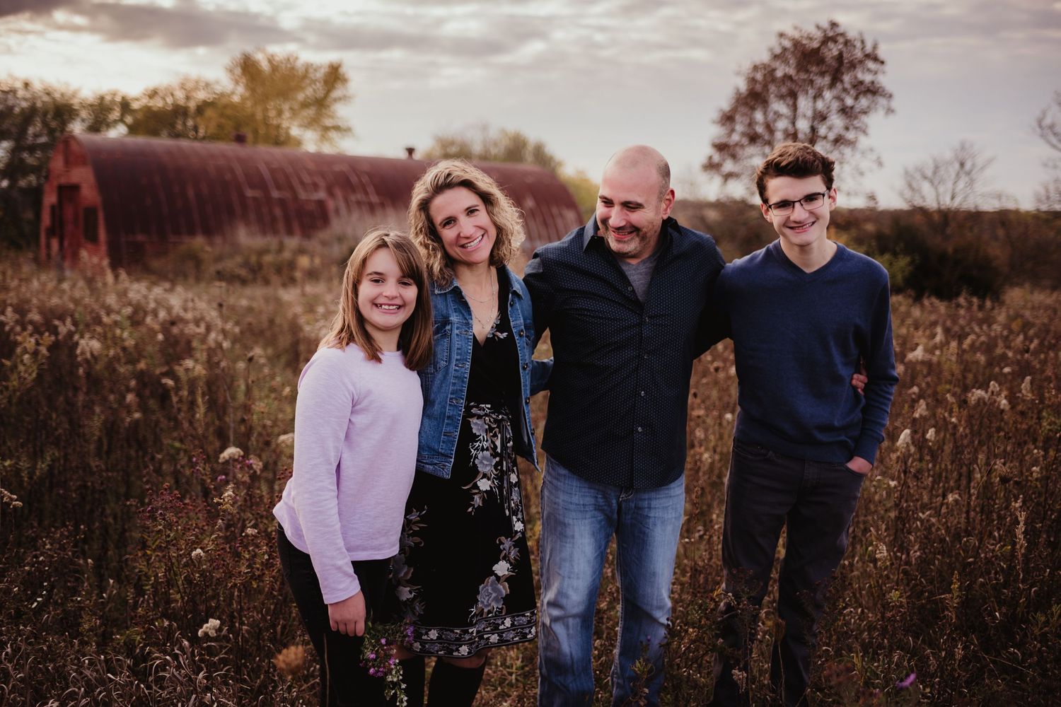 A family of four poses together in a fall field at sunset against a rustic red barn wall.