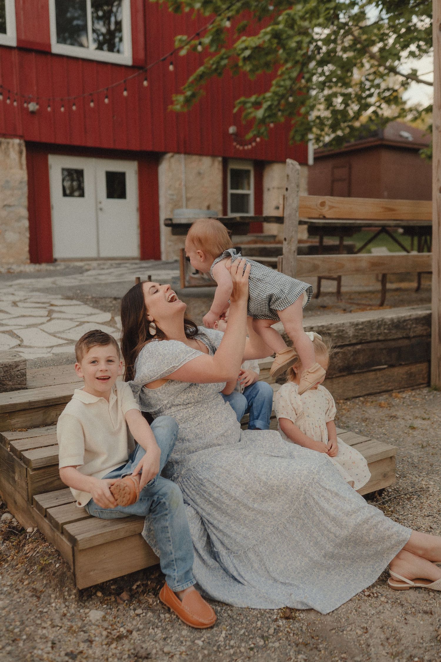 Joyful mother laughing while lifting baby in air, seated on wooden steps with two children beside a rustic red barn.