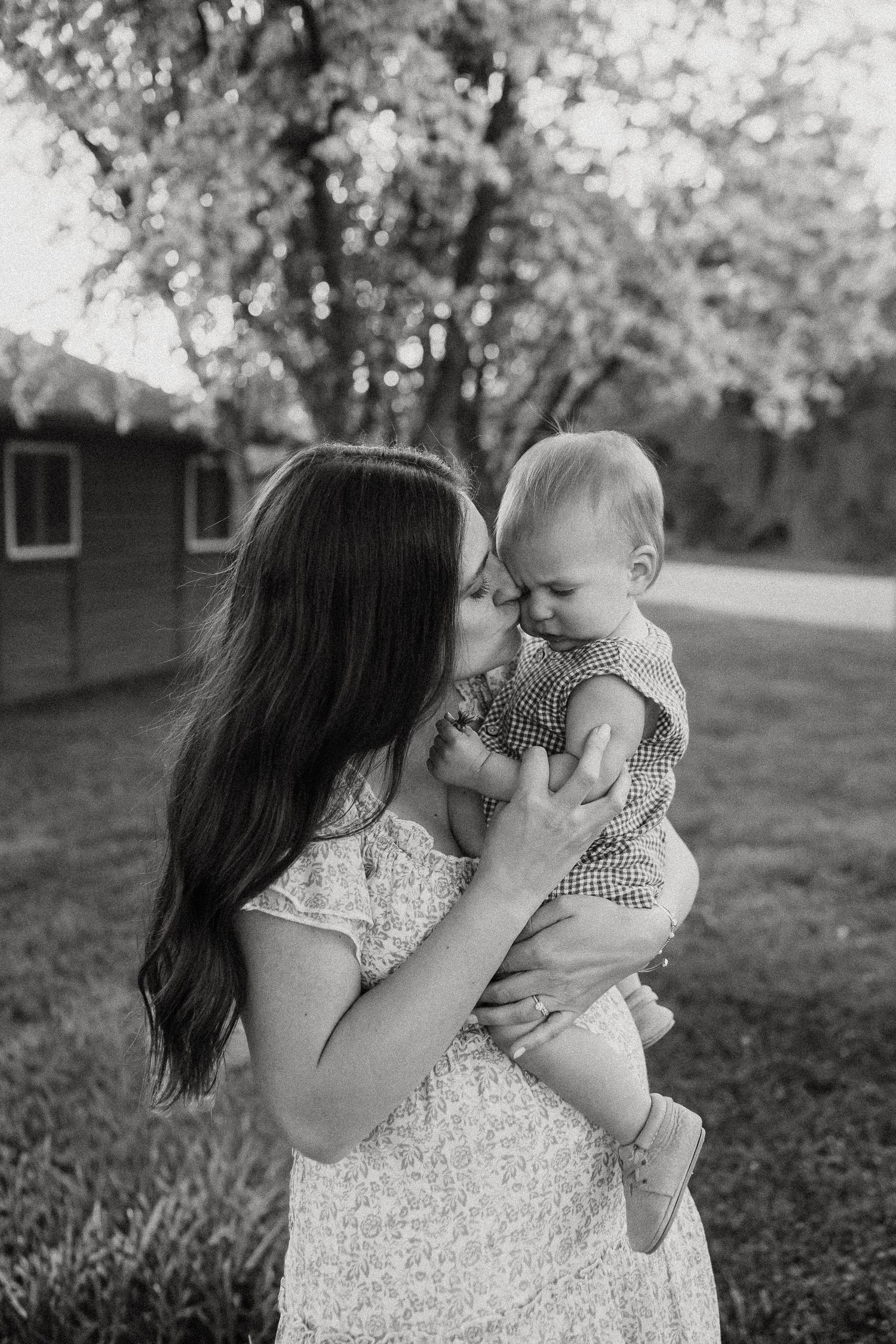 Black and white photo of mother kissing her baby boy tenderly while holding him outdoors.