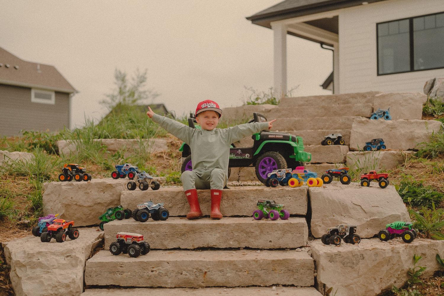 Happy toddler in red boots posing with monster truck toy collection on stone steps.