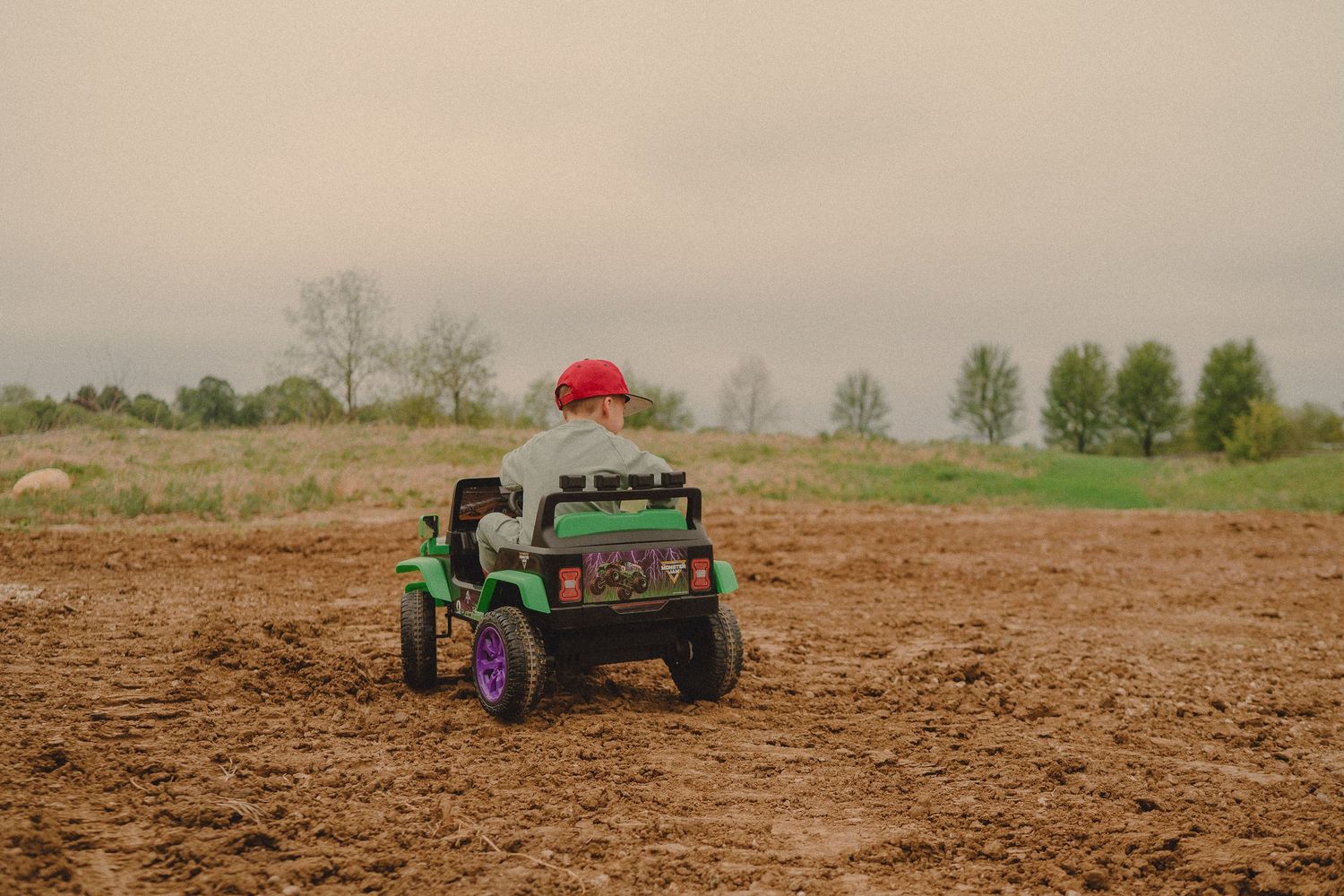 Child riding a green toy jeep on a dirt field with trees in the background on a cloudy day.
