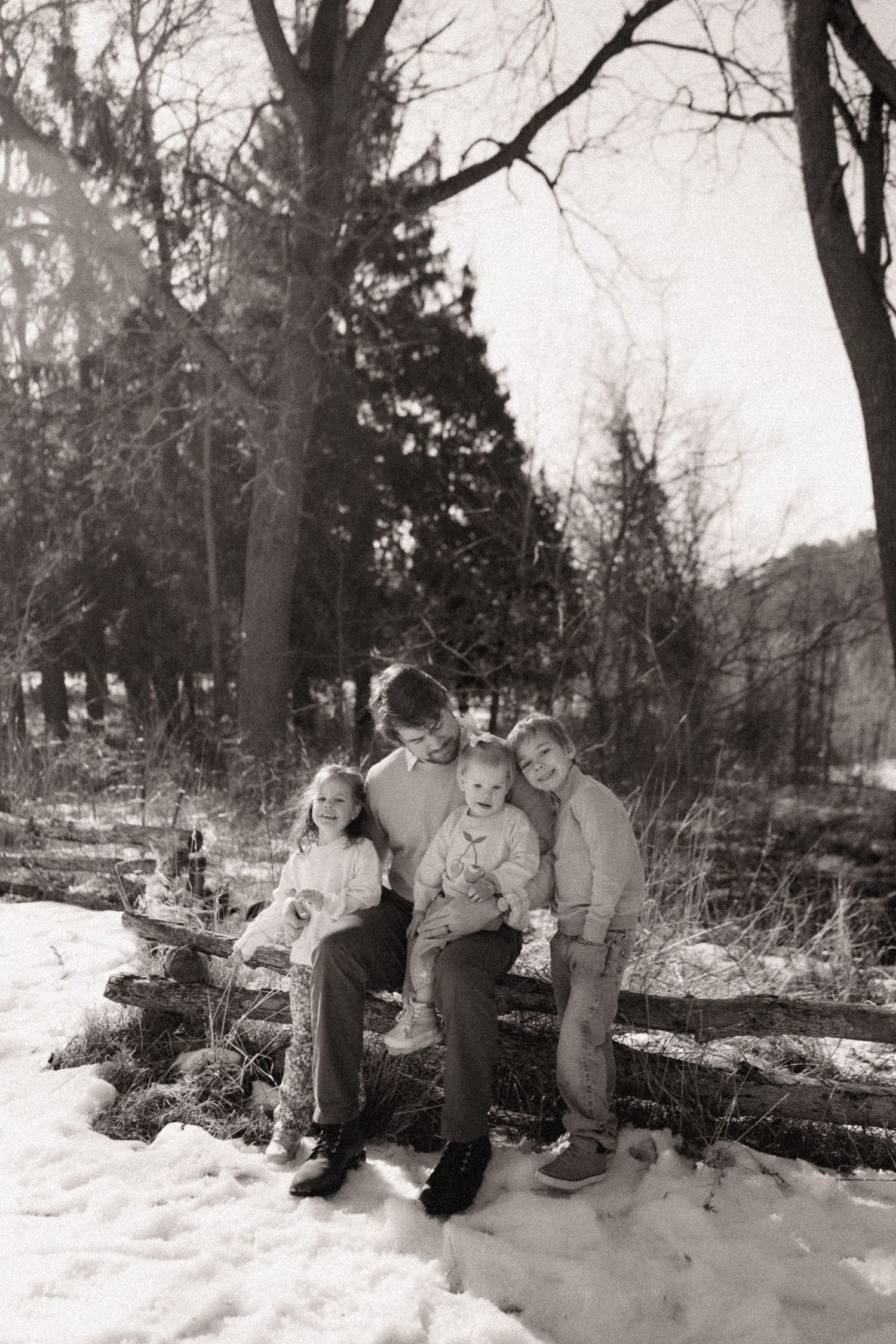 A family group sits together on snowy logs in a winter forest scene captured in black and white.