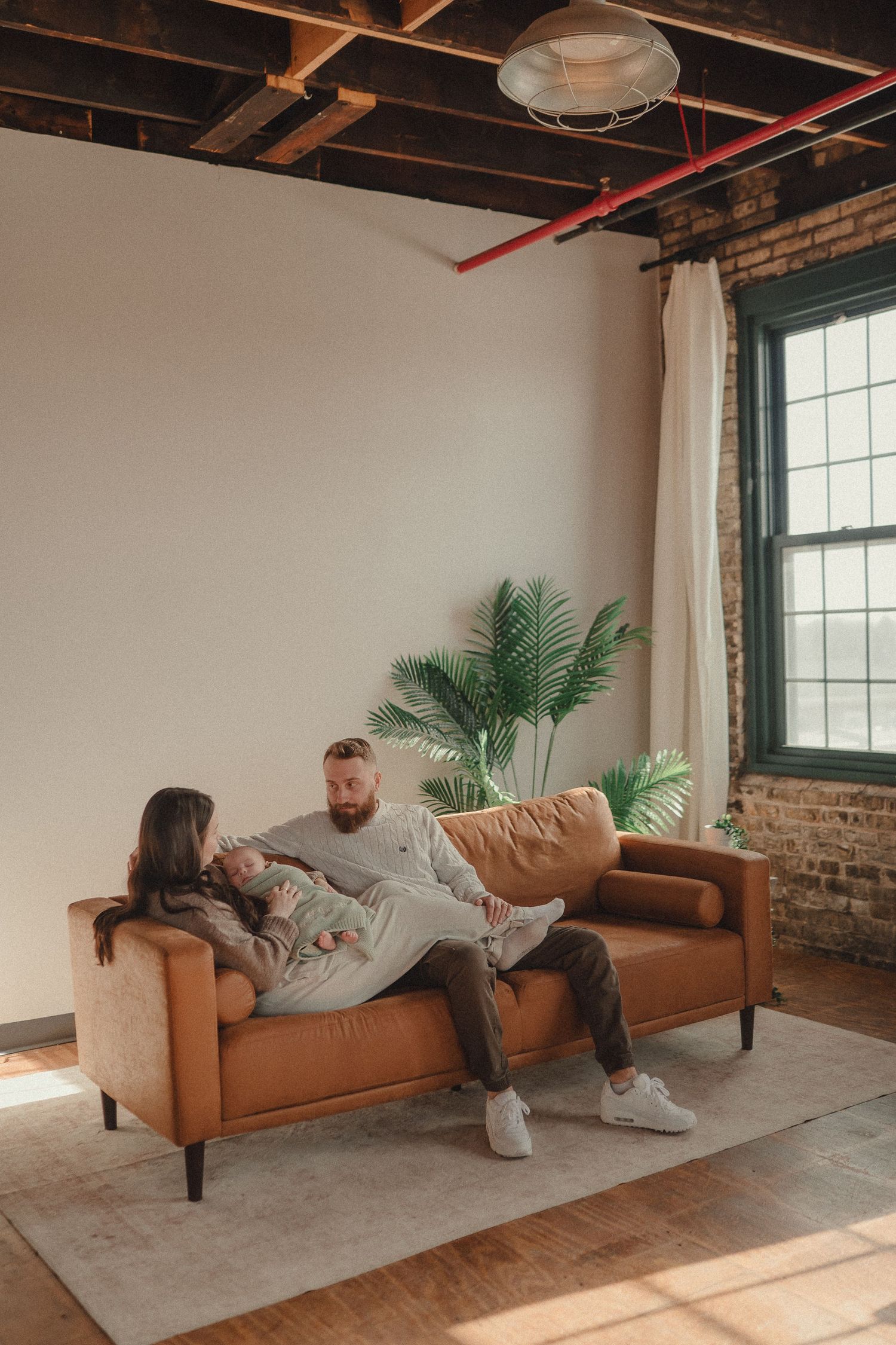 A family relaxes on a tan leather couch in an industrial style room with exposed brick and palm plants.