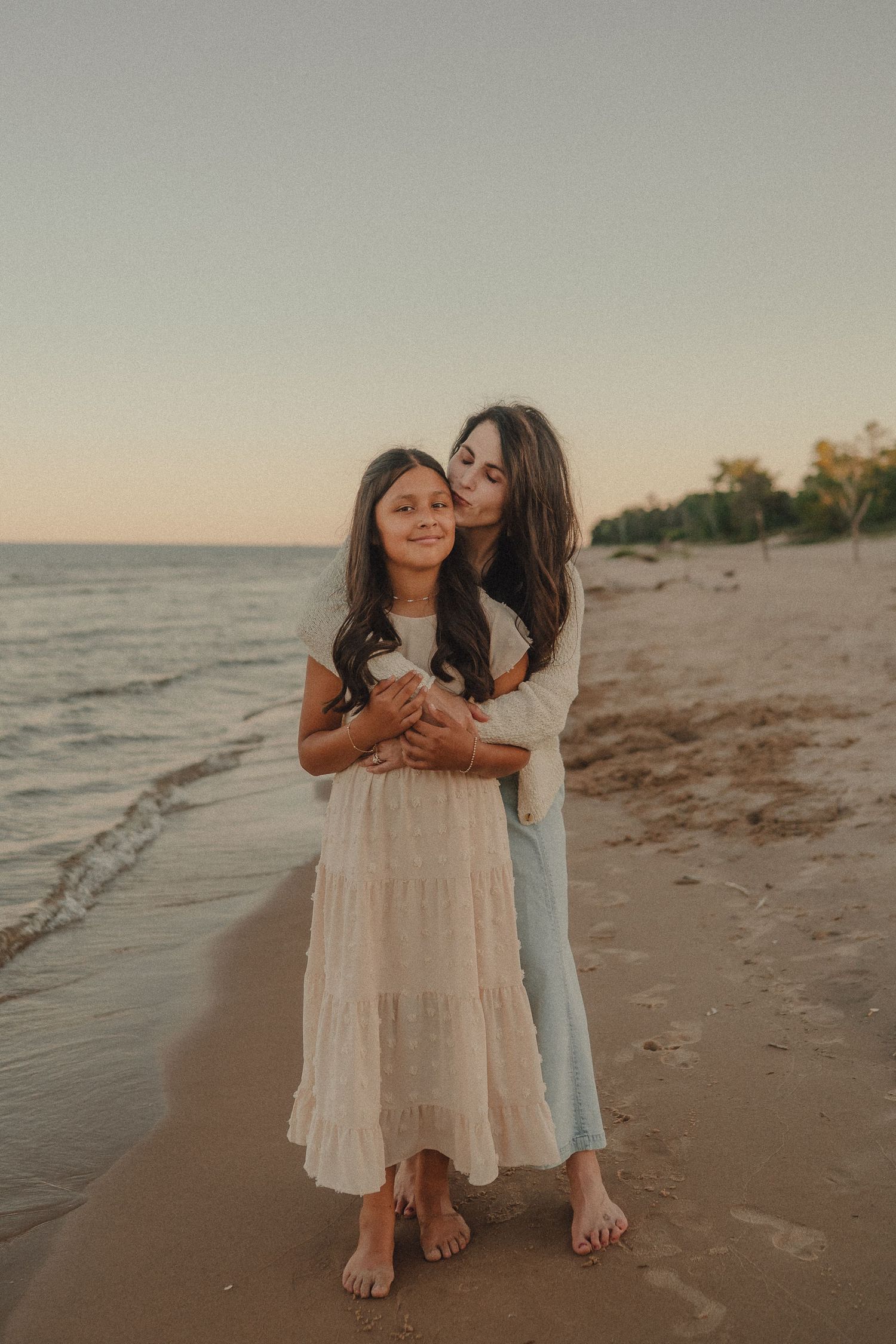 Mother kissing daughter on cheek while embracing on sandy beach at sunset.