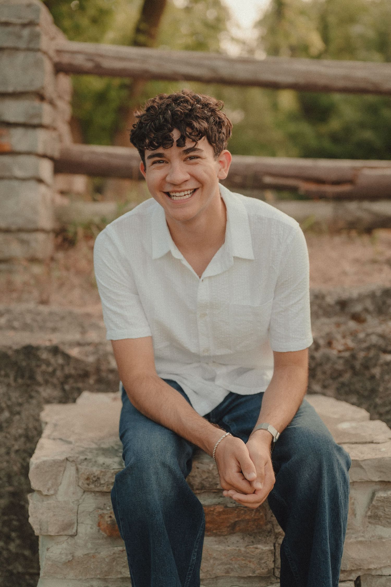 Smiling young man with curly hair sitting on stone wall wearing white shirt and jeans outdoors.