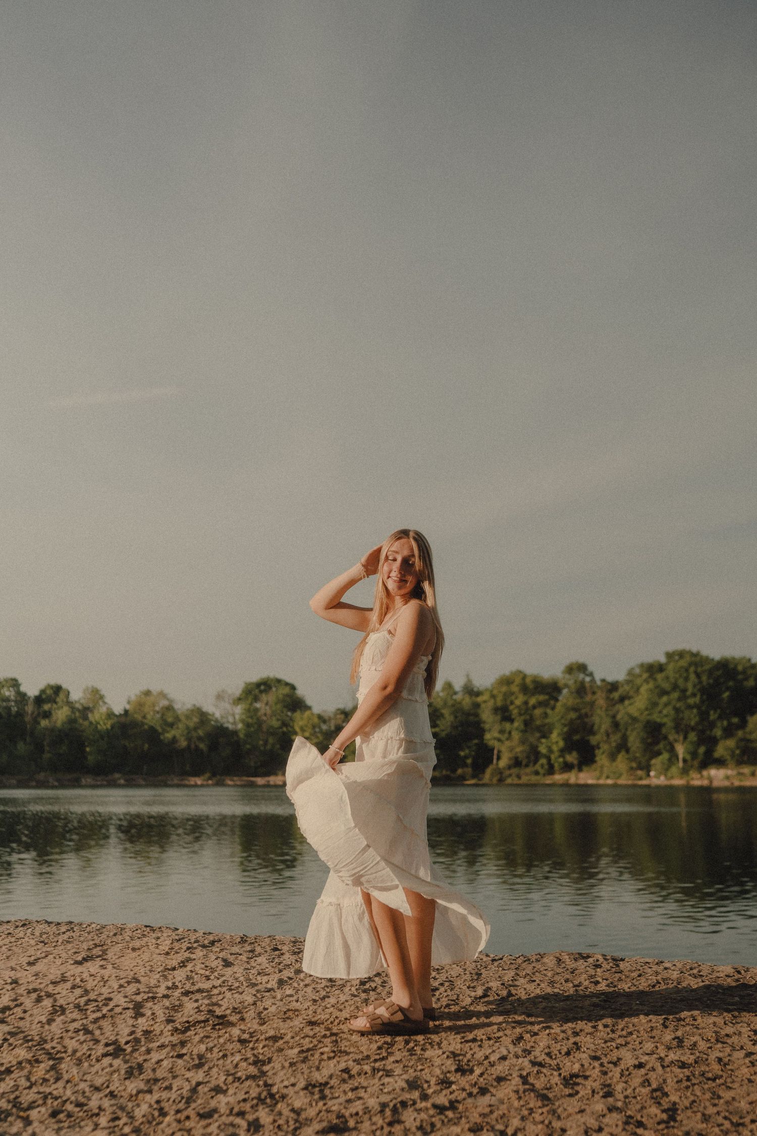 A woman in a white flowing dress stands on a sandy lakeside shore, smiling with trees and water in the background.