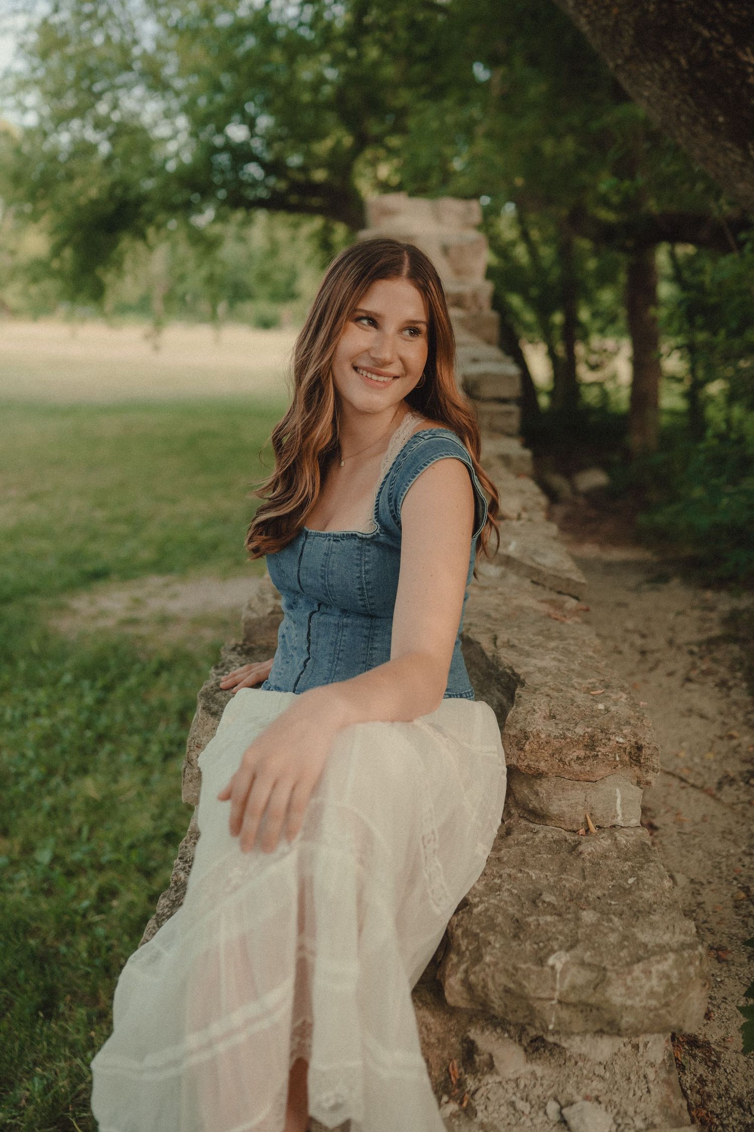 Young woman in denim top and white skirt sits smiling on a stone wall outdoors.