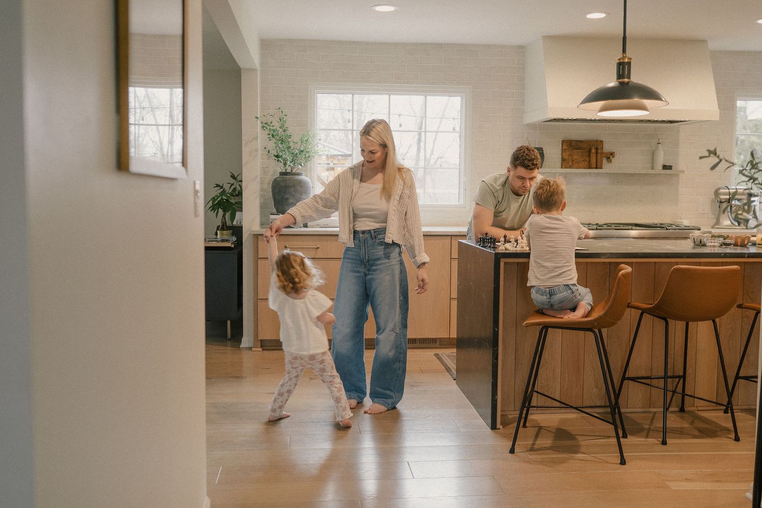 A mother dances with her toddler in a kitchen while father and child play at the counter.