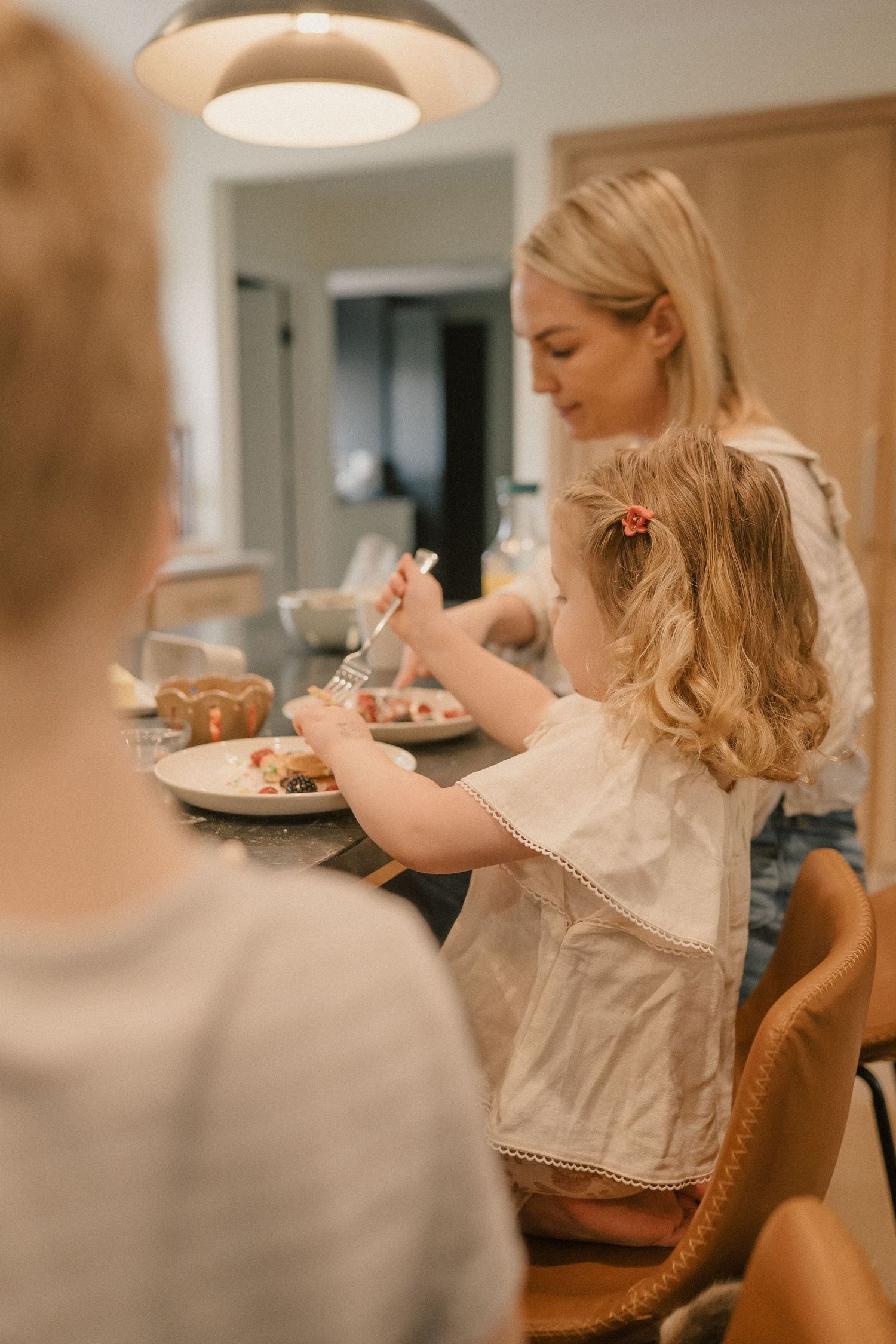 A mother helps her young daughter eat at a kitchen table under warm pendant lighting.