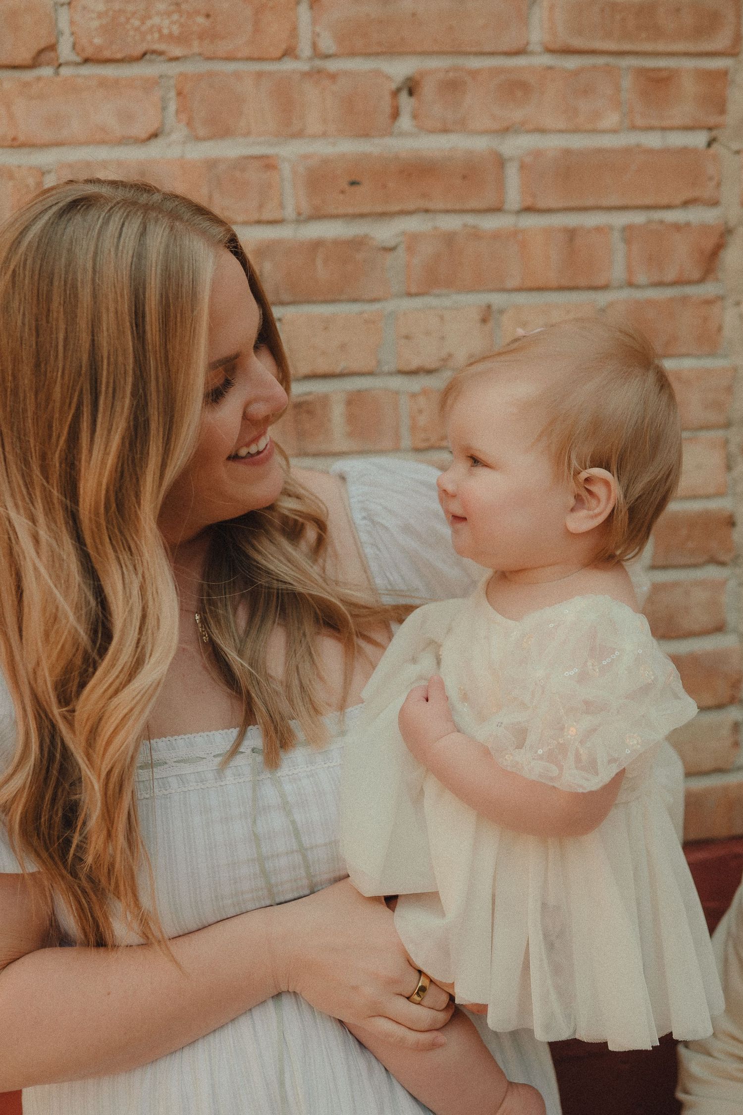 A loving mother gazes at her toddler daughter in a white lace dress during an outdoor portrait session.