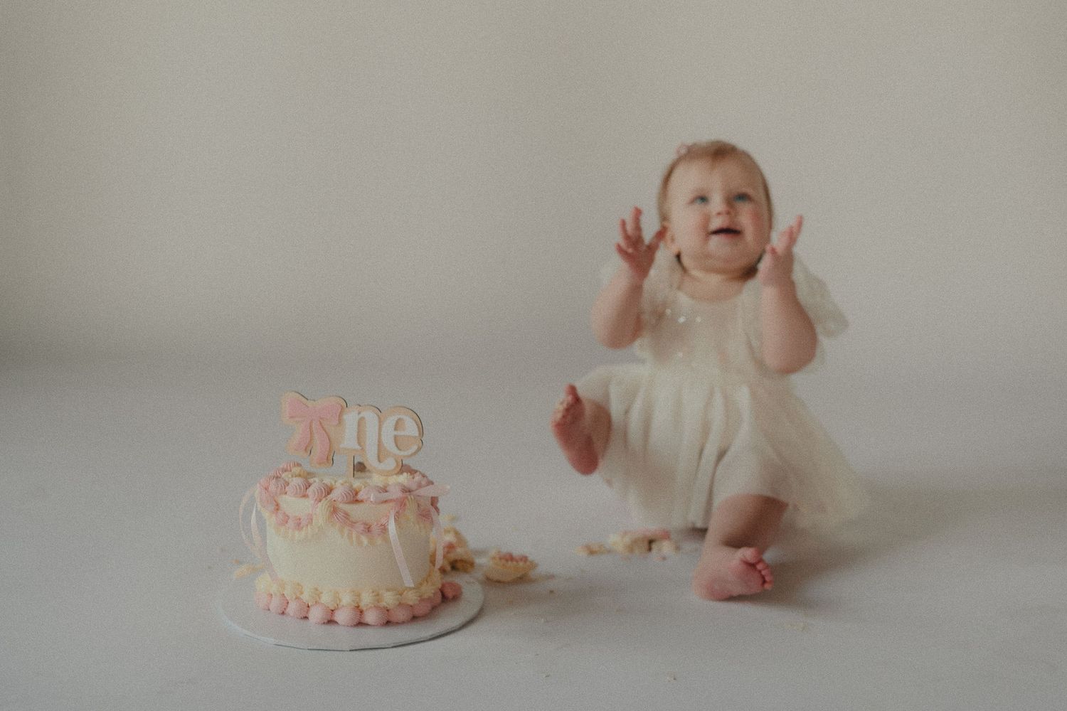 Joyful baby girl in white dress claps frosting-covered hands next to her pink first birthday smash cake.
