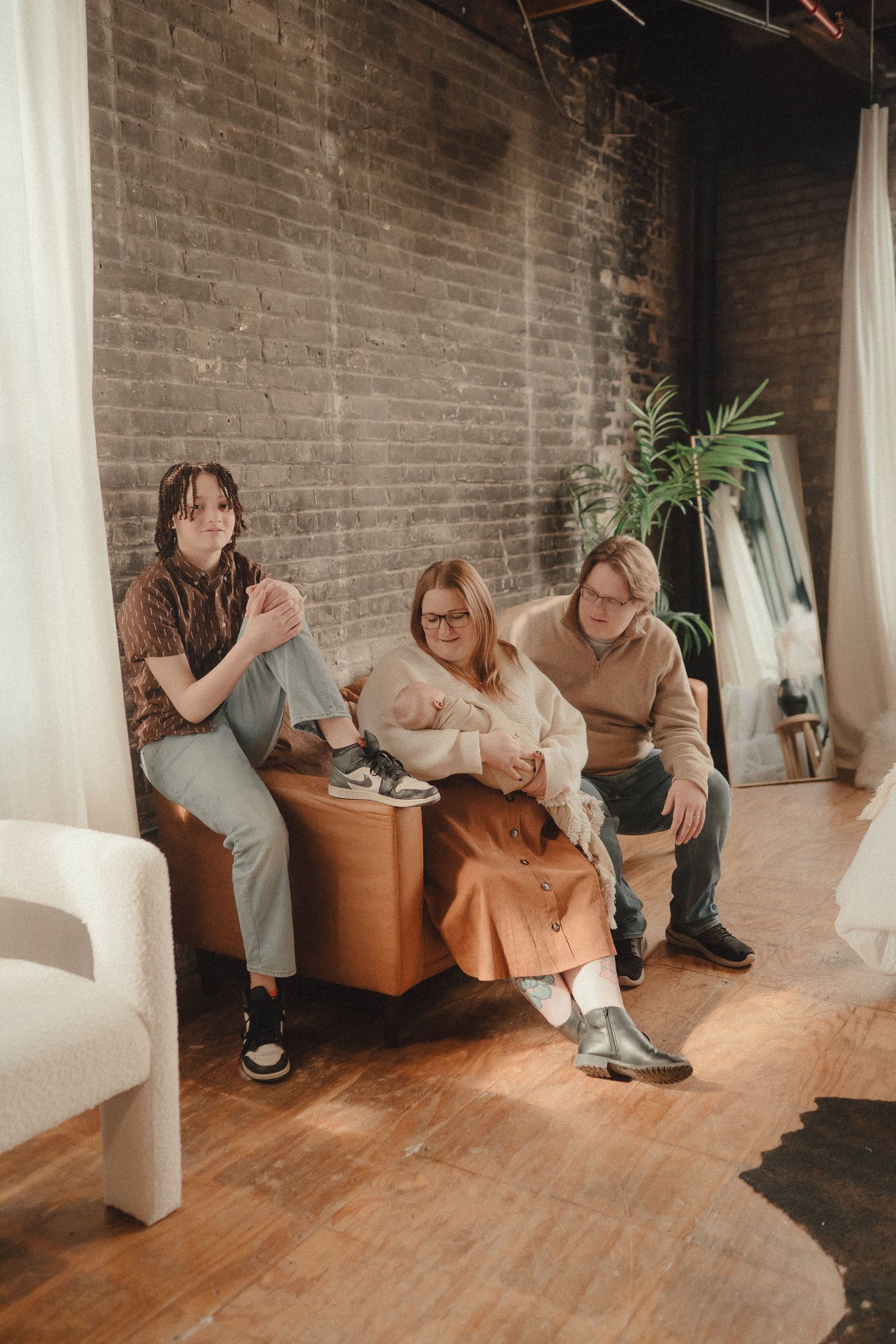 Family of three sits together on brown leather ottoman against exposed brick wall in modern loft.
