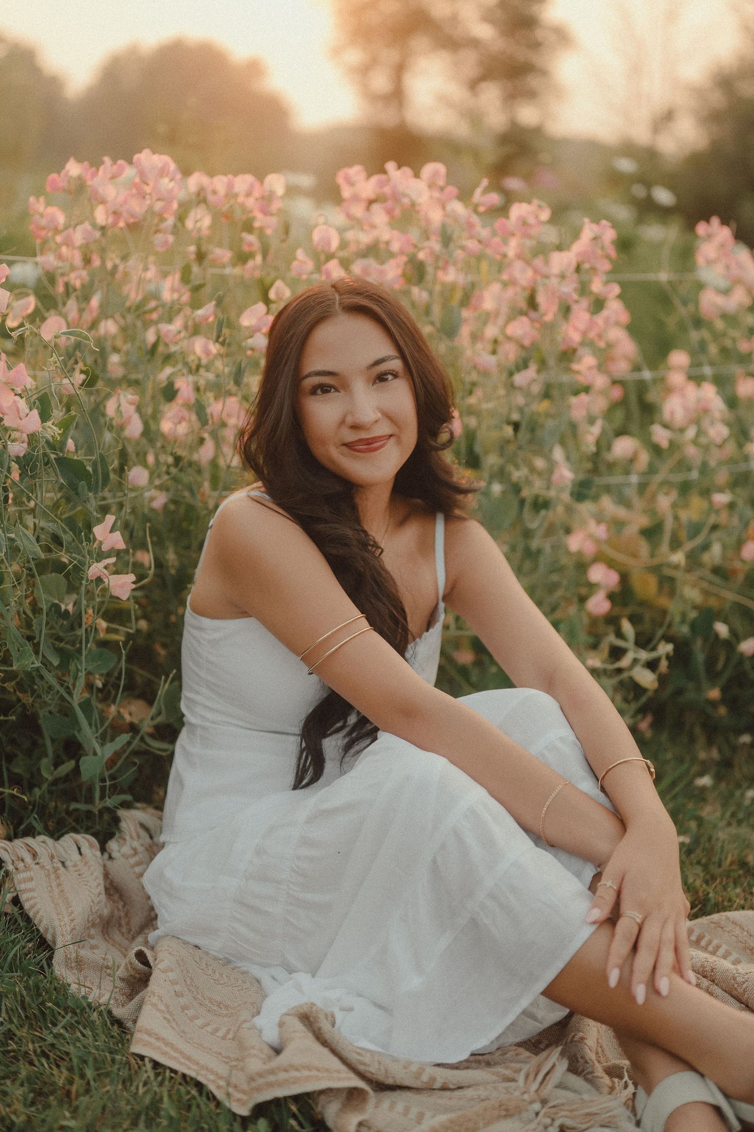 Young woman in white dress sitting among pink wildflowers at golden hour.
