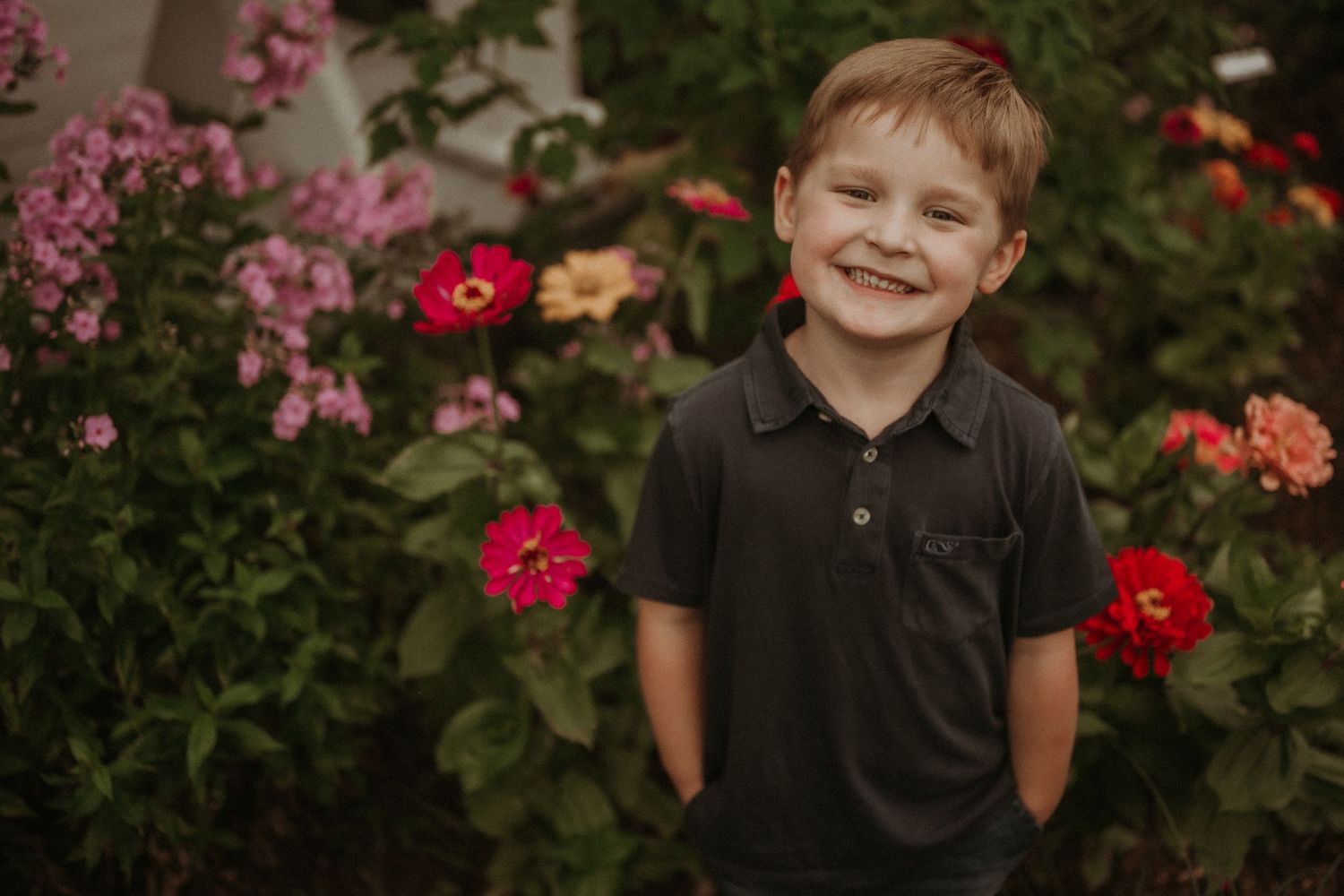 Young child in dark polo shirt smiling brightly against backdrop of red and pink flowers.