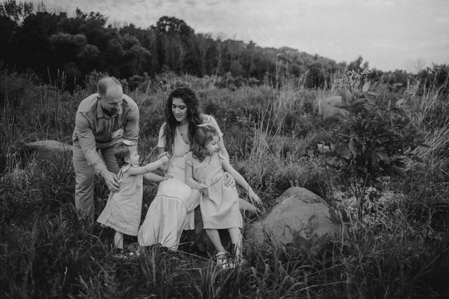 A black and white candid family photograph taken in a rustic outdoor setting with tall grass.