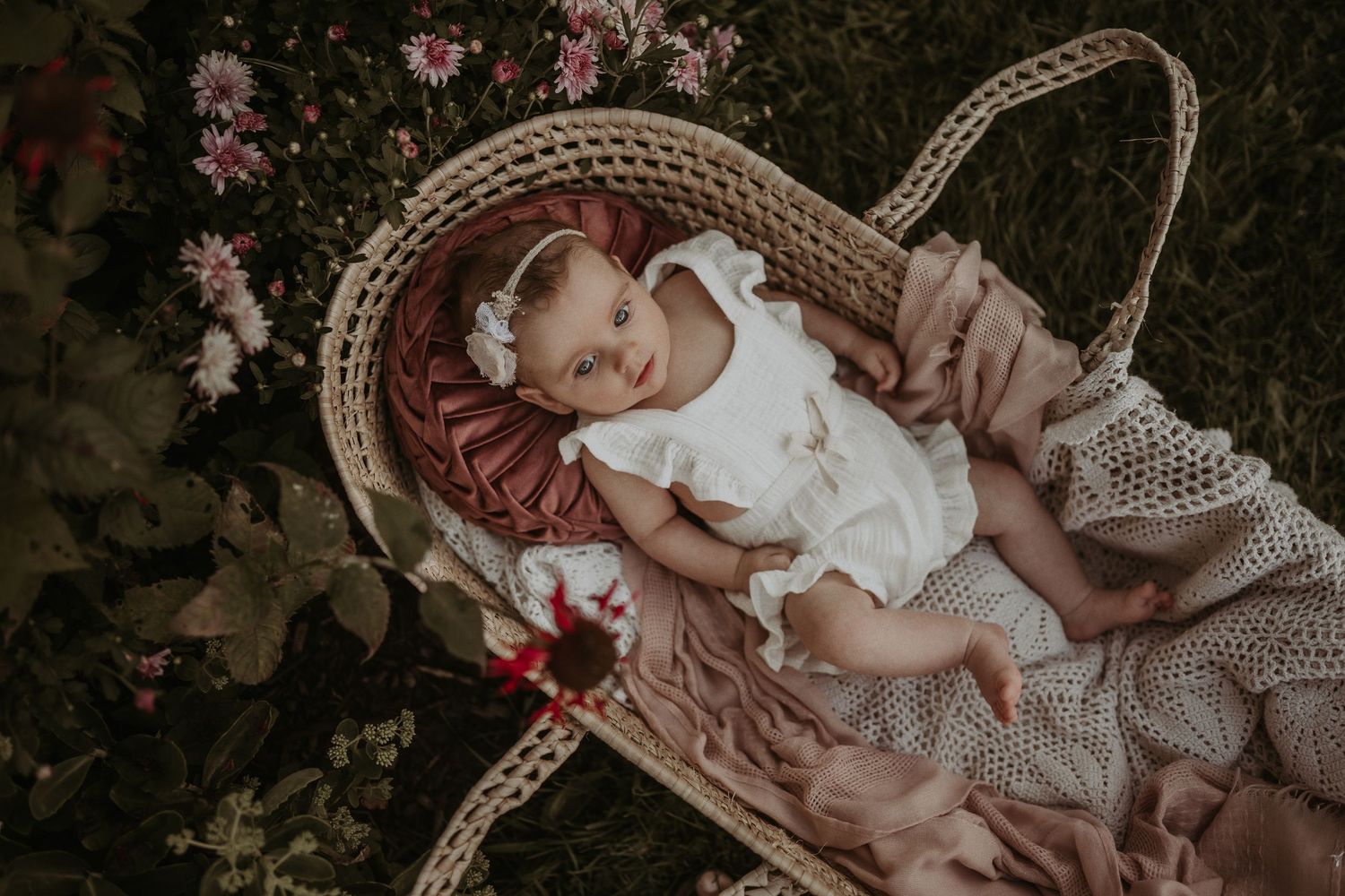 A sleeping baby rests in a woven basket surrounded by pink blossoms.