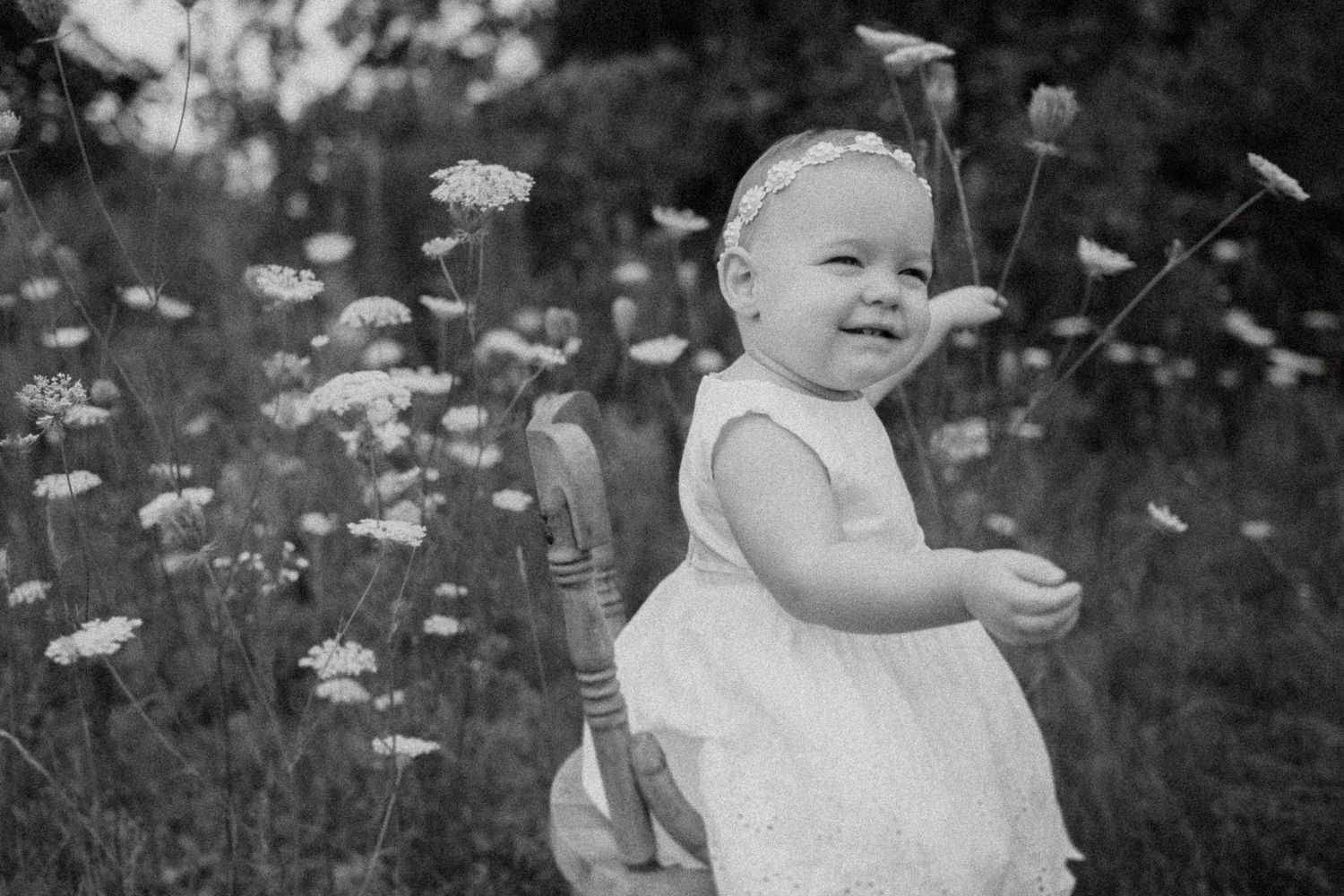 Black and white photo of baby in white dress standing among wildflowers.