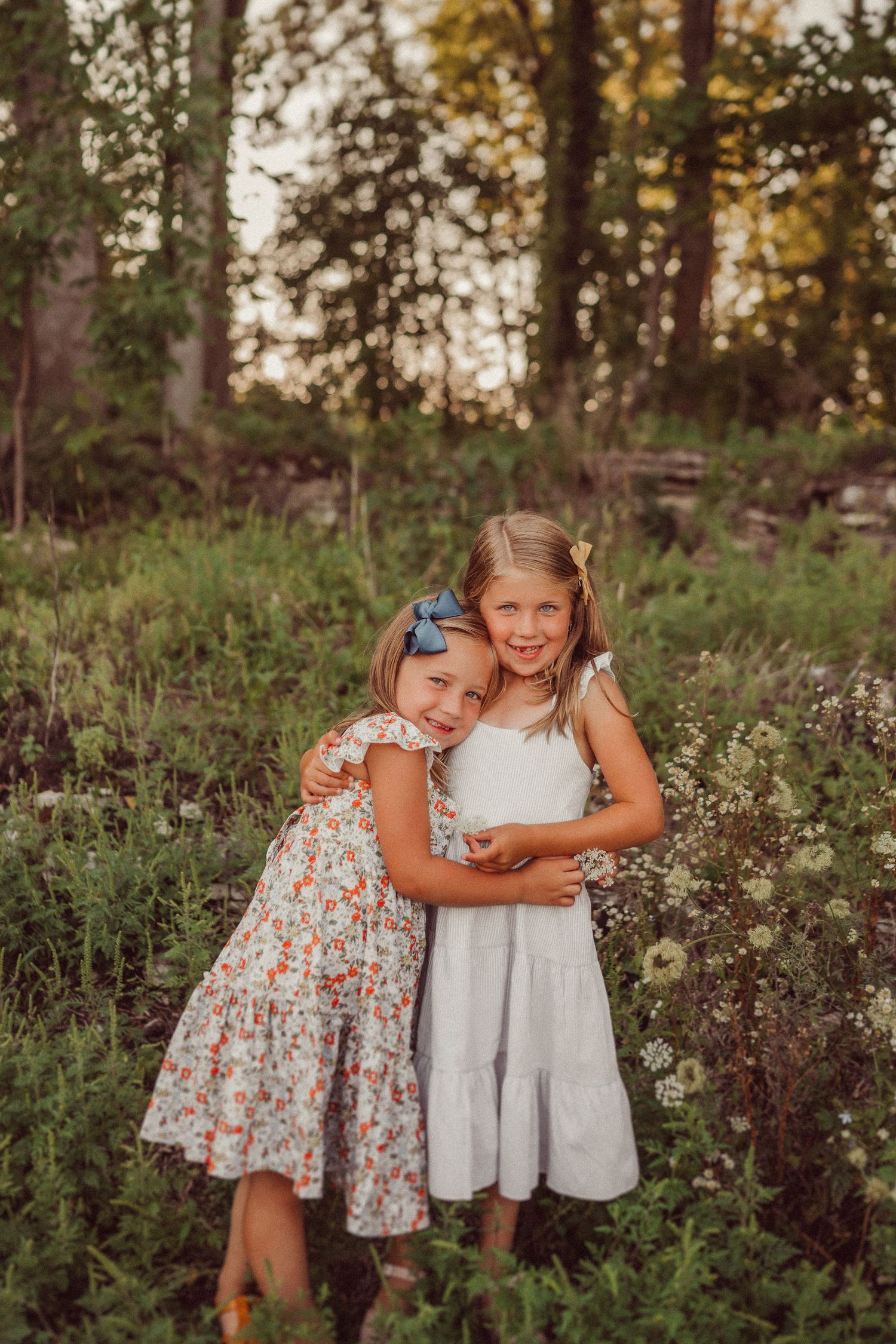 Two young children in summer dresses embrace while standing in a field of wildflowers at sunset.