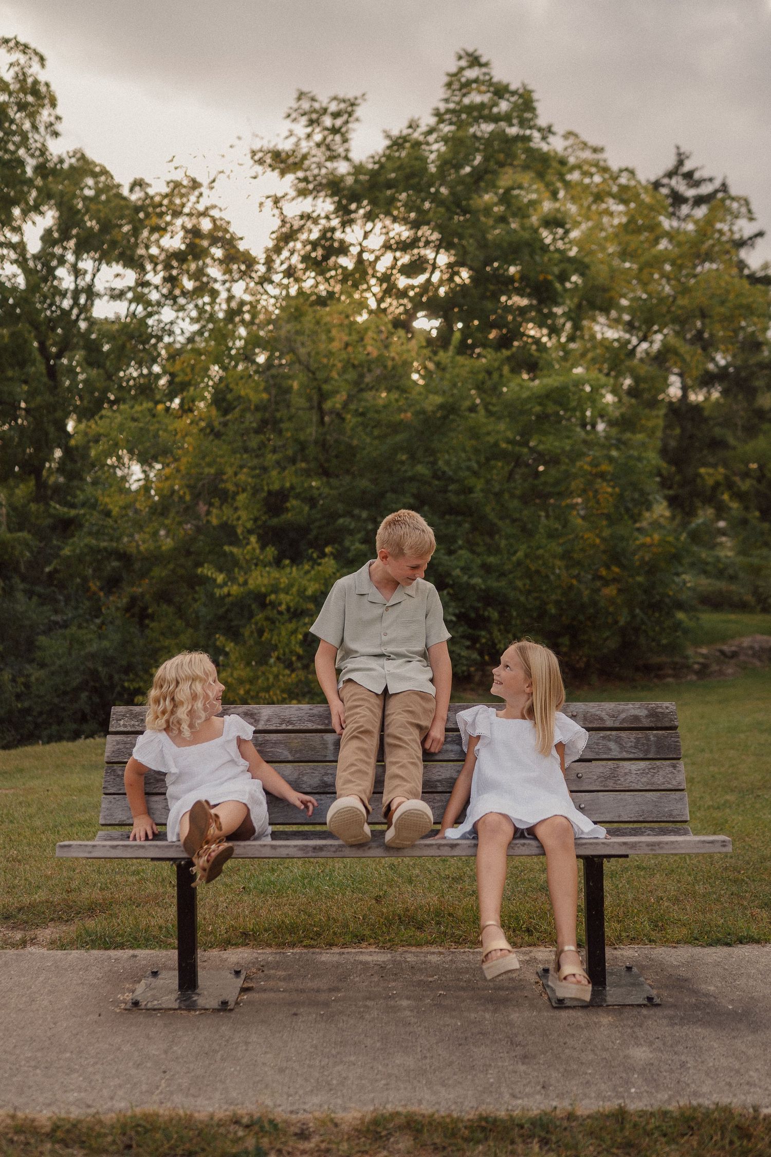 Three children sit together on a wooden park bench at sunset surrounded by green trees in the background.