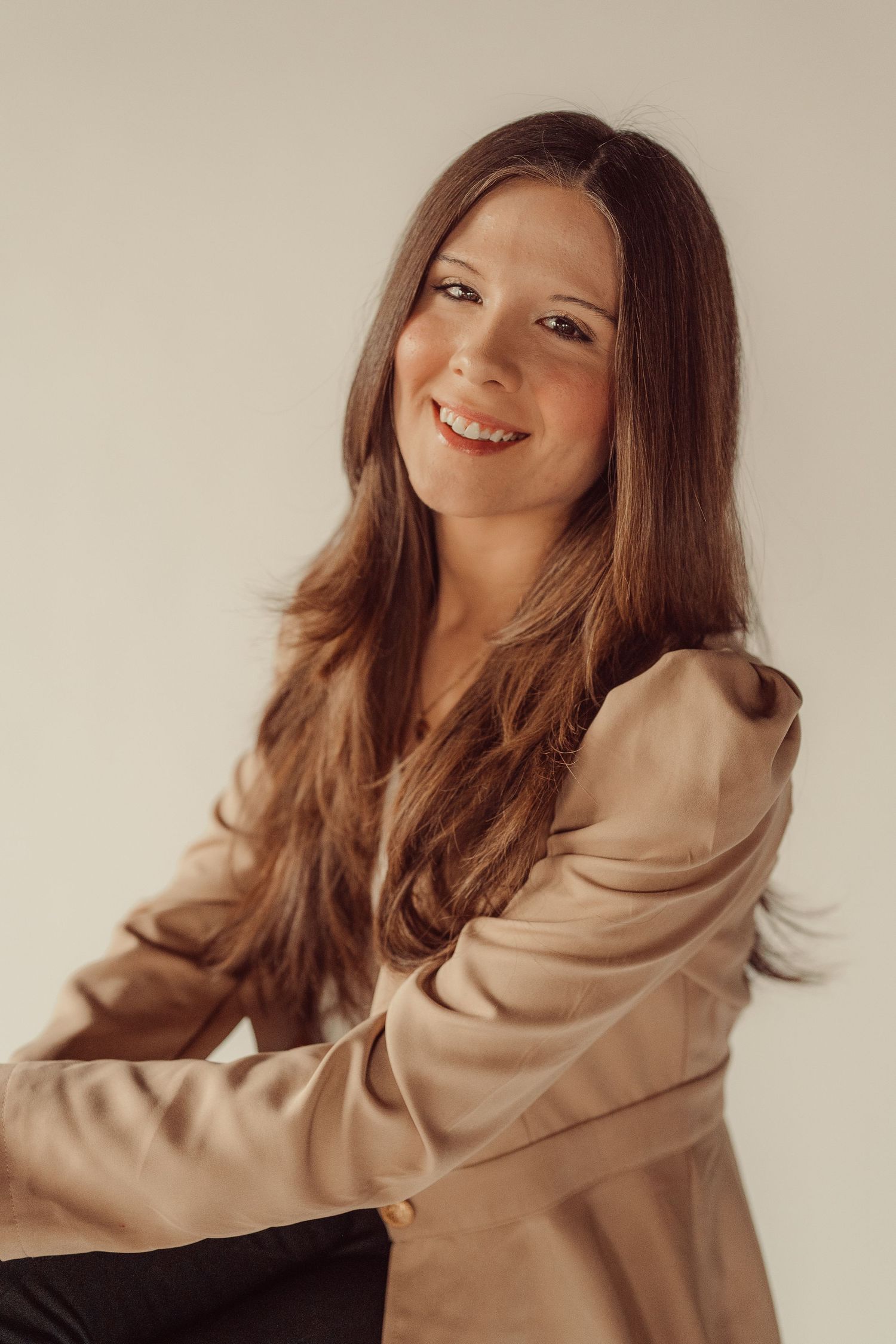 Professional portrait of woman in beige blazer smiling warmly against neutral background.
