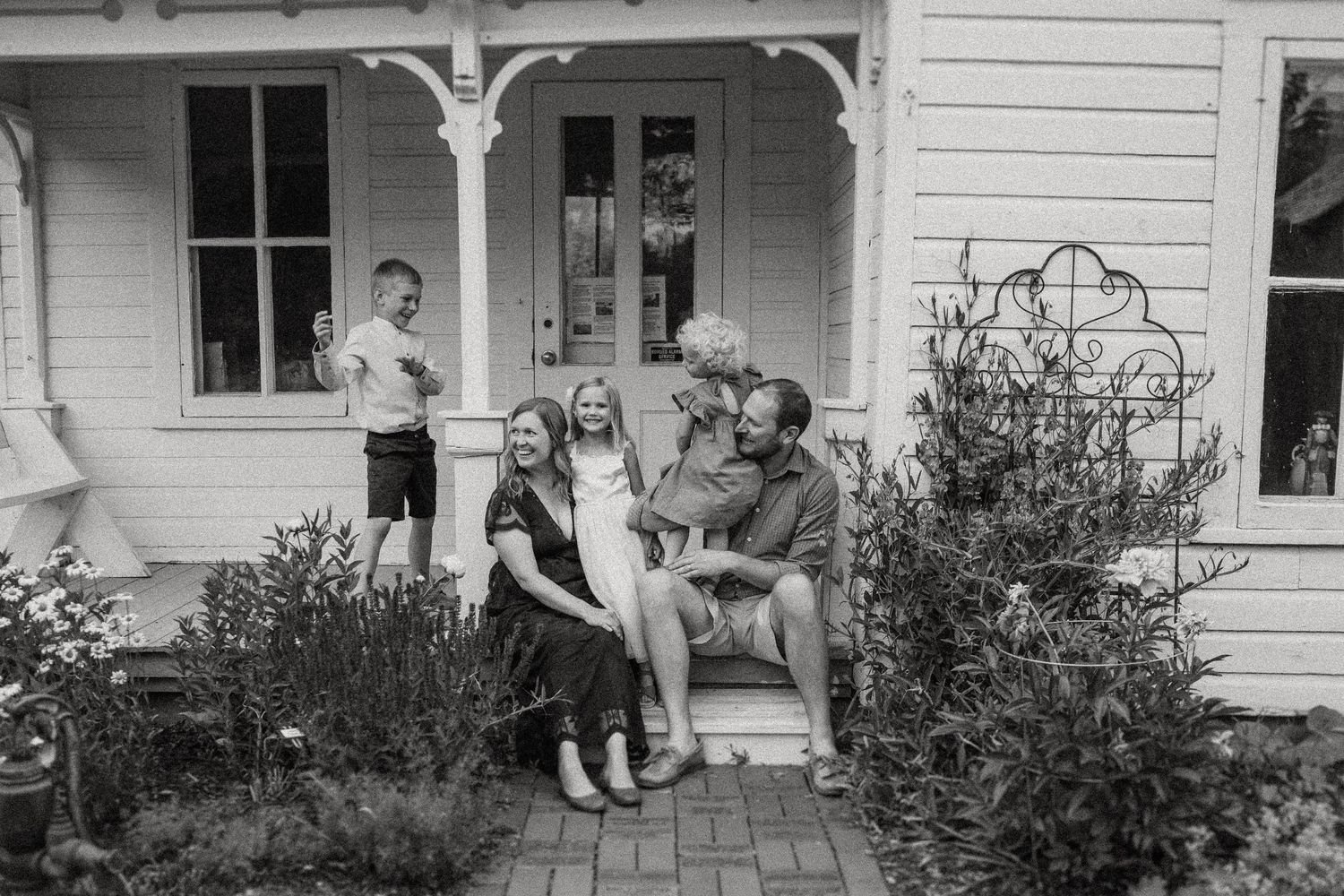 Group gathered on white farmhouse porch with blooming garden and decorative trellis in foreground.