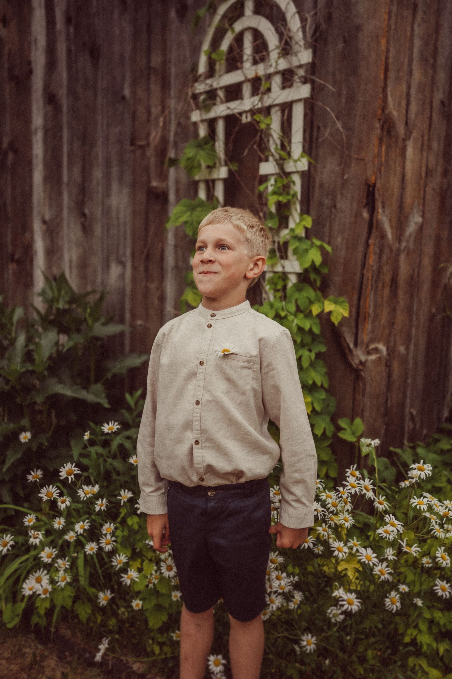 A child in a beige button-down shirt and shorts stands by a rustic wooden fence with climbing vines.