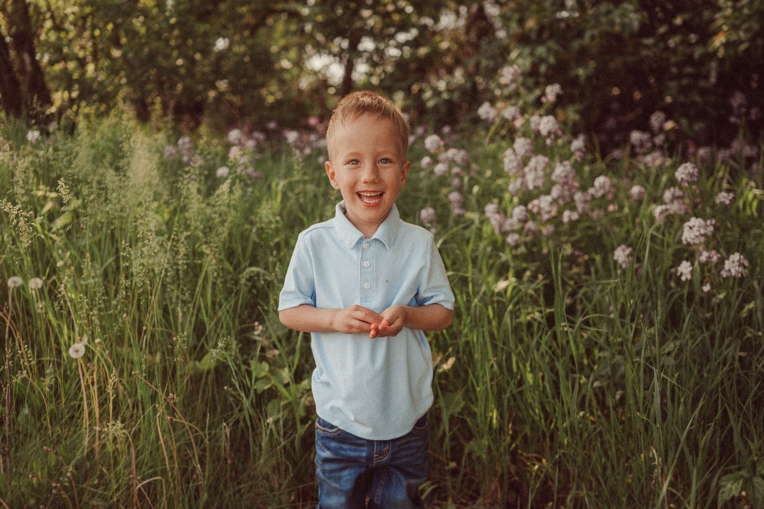 A cheerful child in a light blue shirt stands among tall grass and blooming flowers.