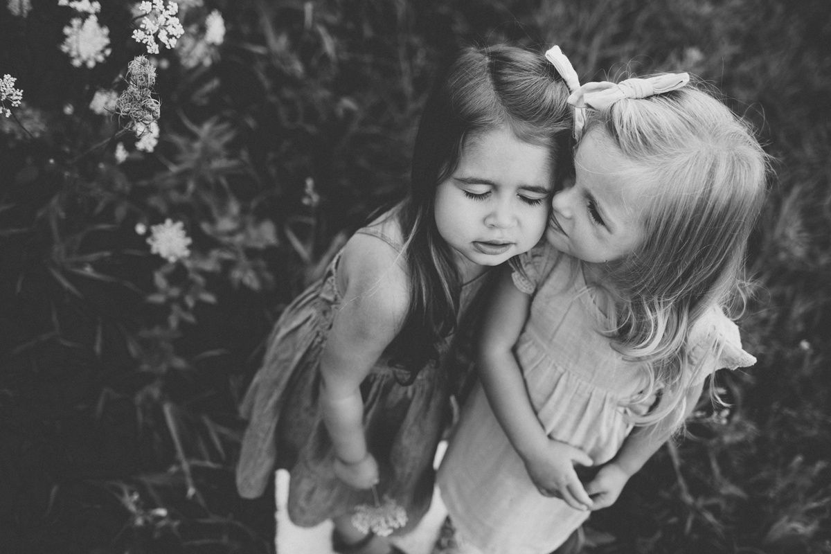 A black and white candid photo of two children embracing outdoors near flowers.