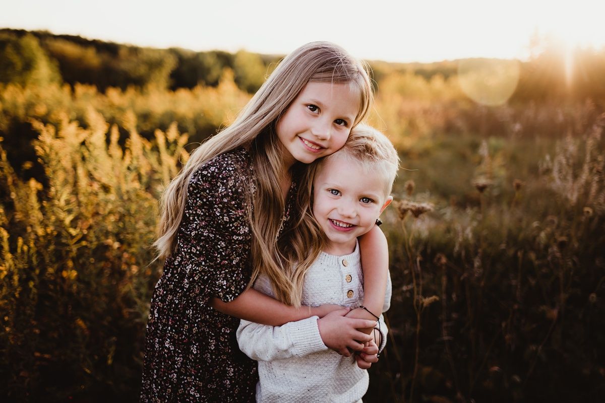 Two siblings embrace while laughing together in sunlit autumn field.