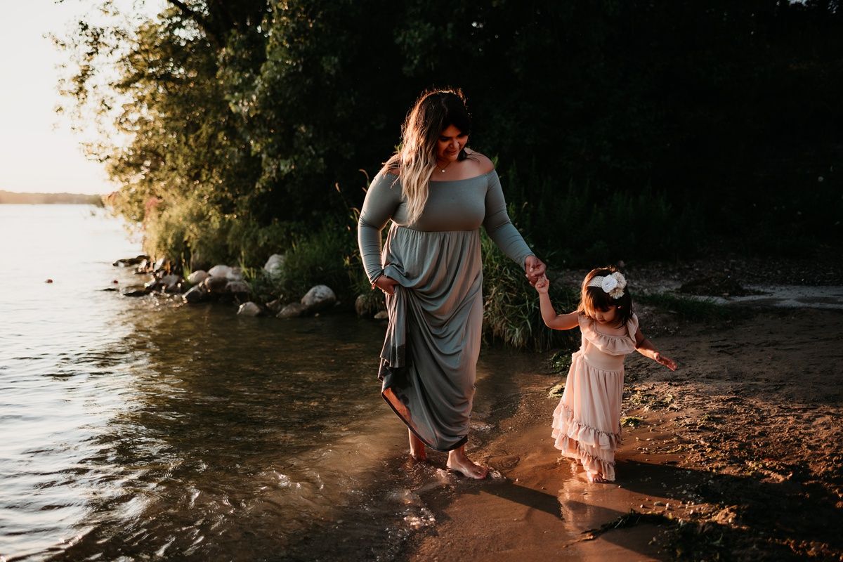 Mother and child holding hands while walking along water's edge at sunset.