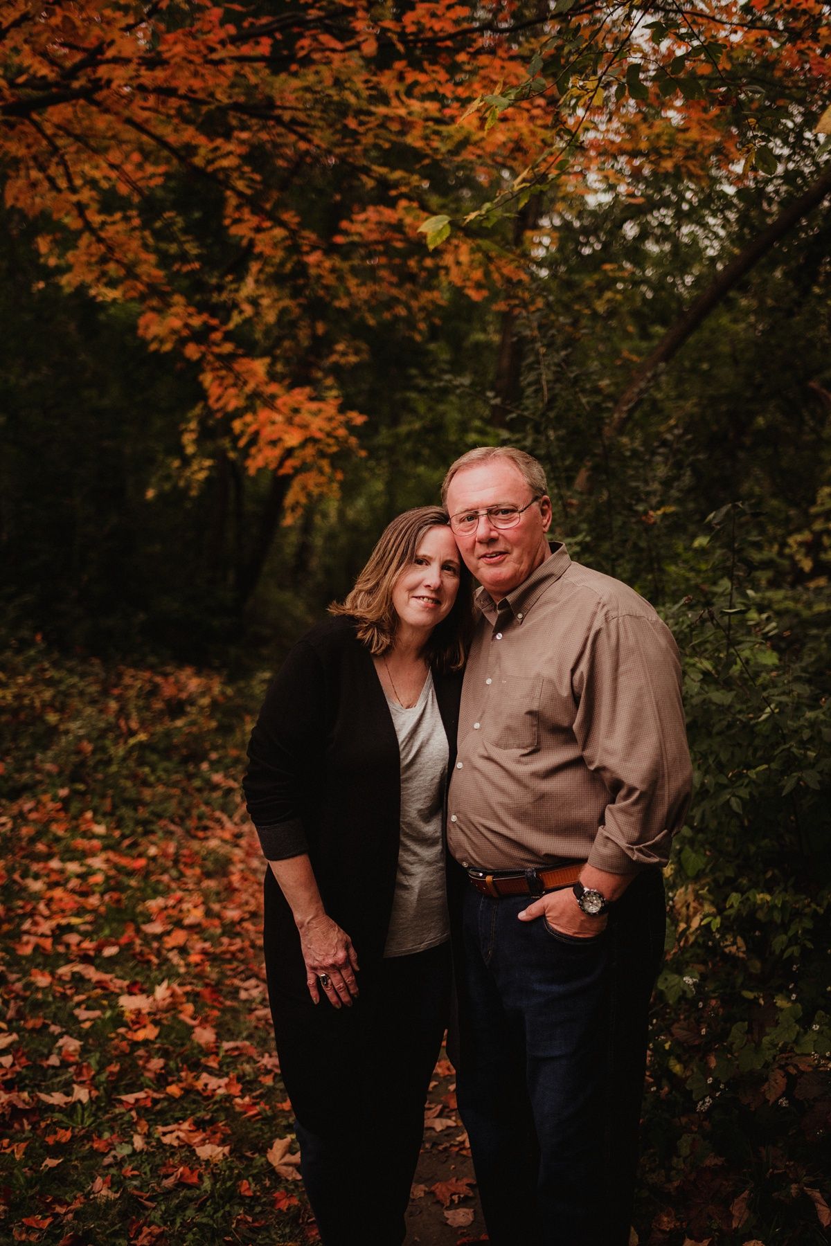 Mature couple poses together in autumn forest with orange leaves in background.