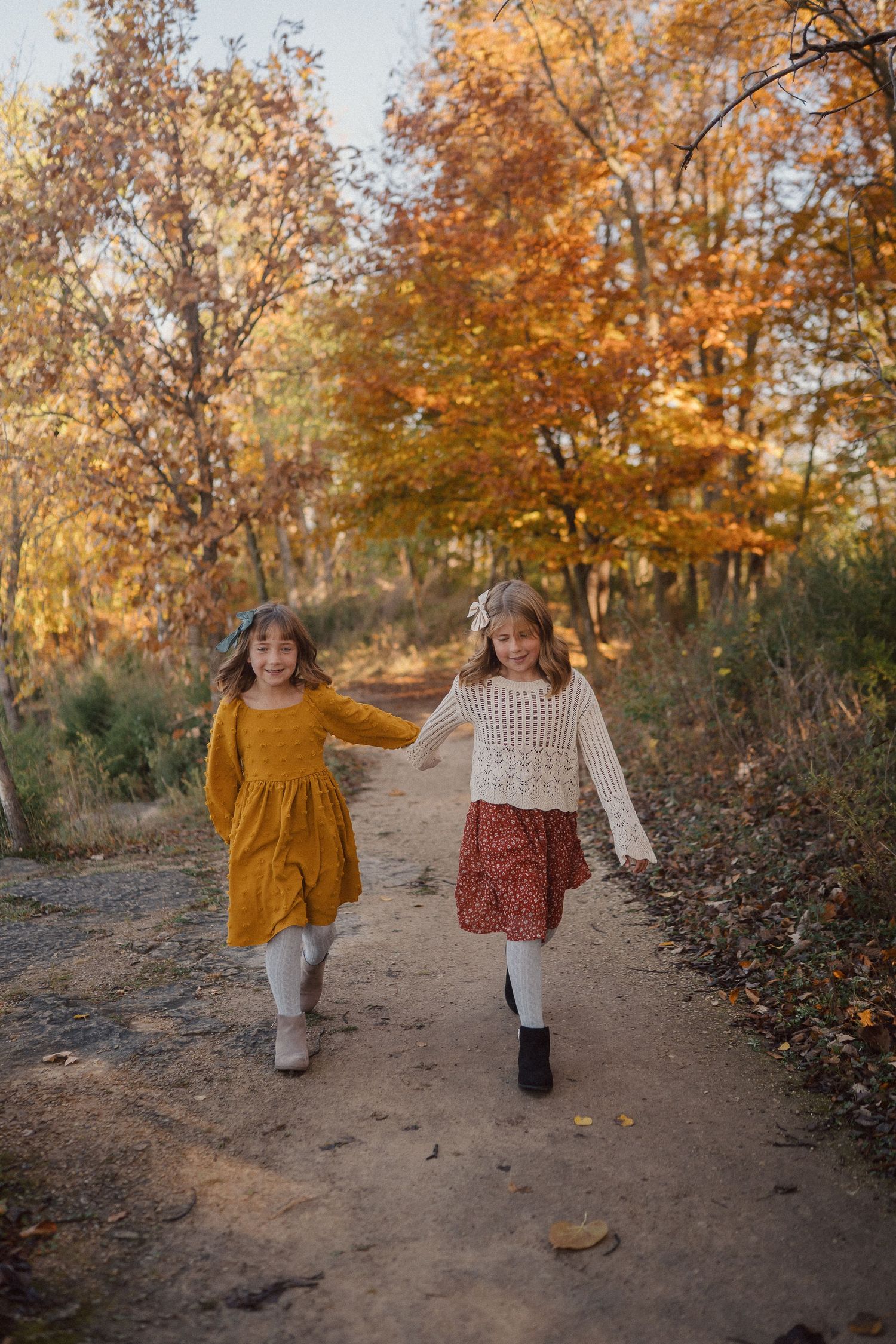 Two children in vintage-style dresses holding hands while walking along an autumn forest path.