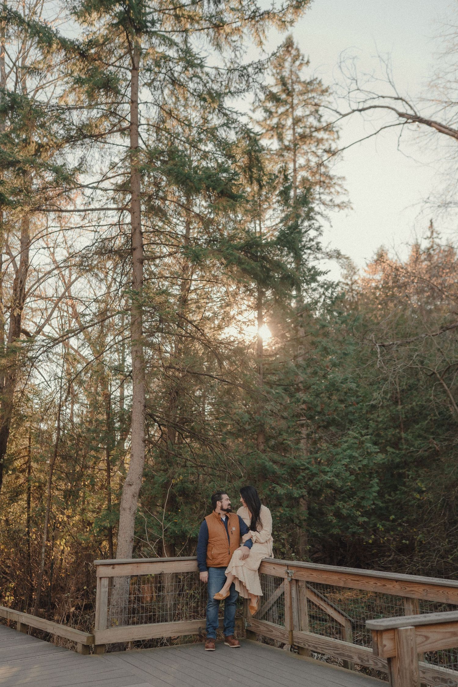 A couple stands on a wooden observation deck surrounded by evergreen trees at sunset.