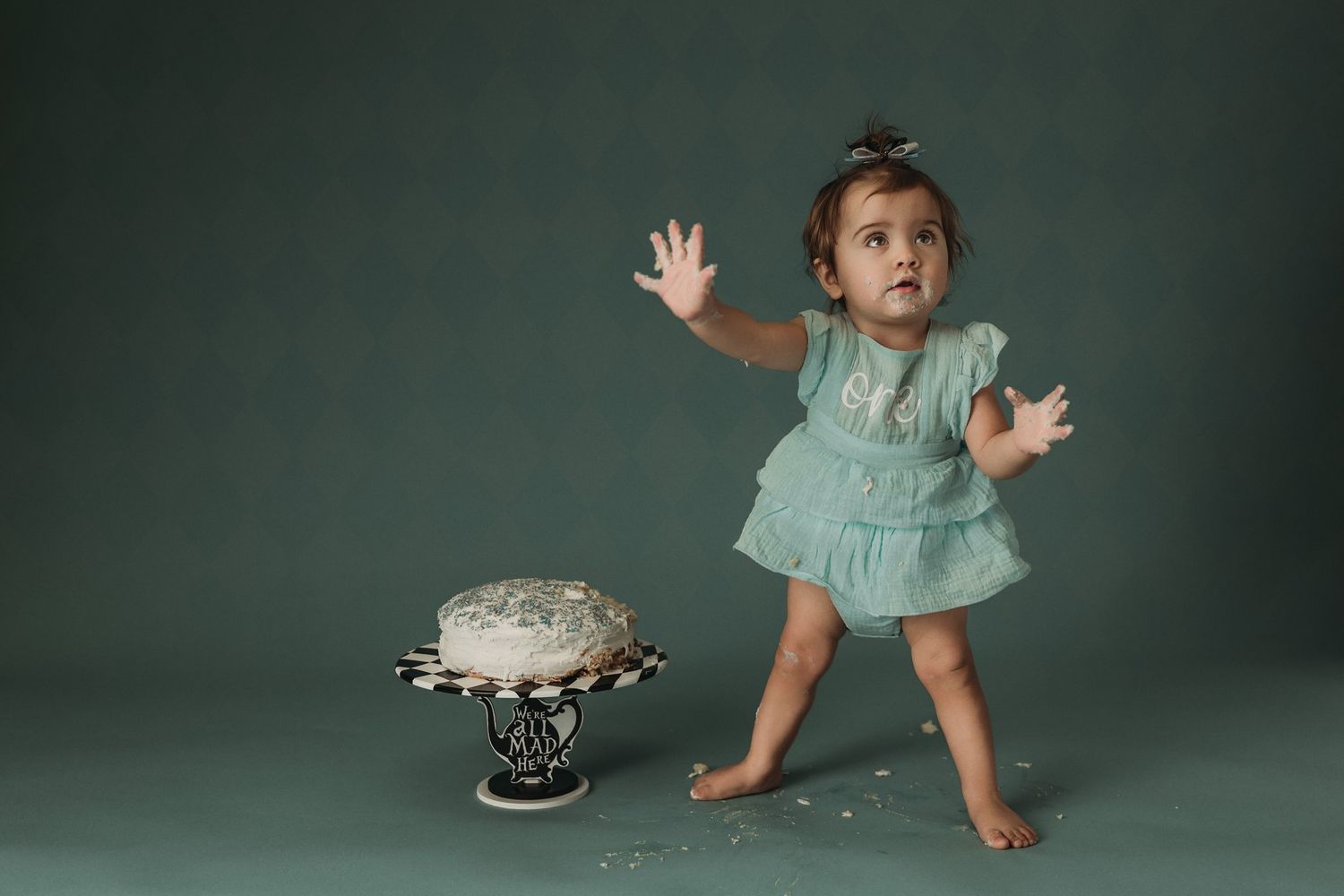 A tiny figure in a mint dress dances next to an ornate cake stand with a white cake.