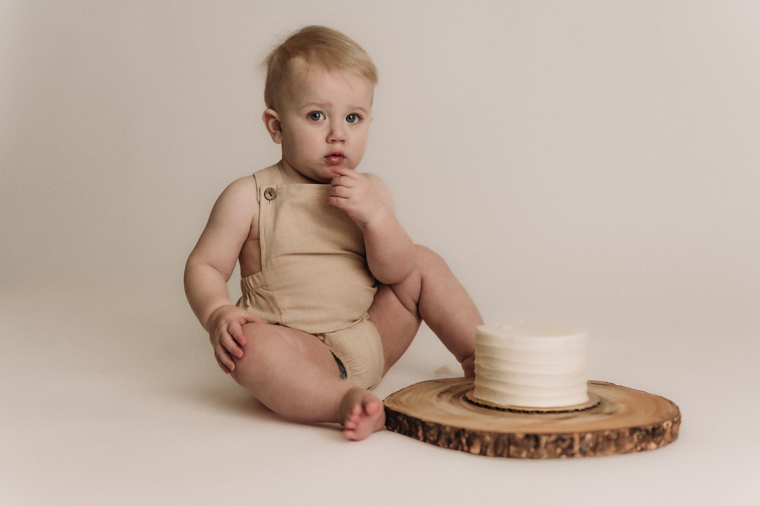 Baby in beige outfit sits beside white cake on rustic wood slice against neutral backdrop.